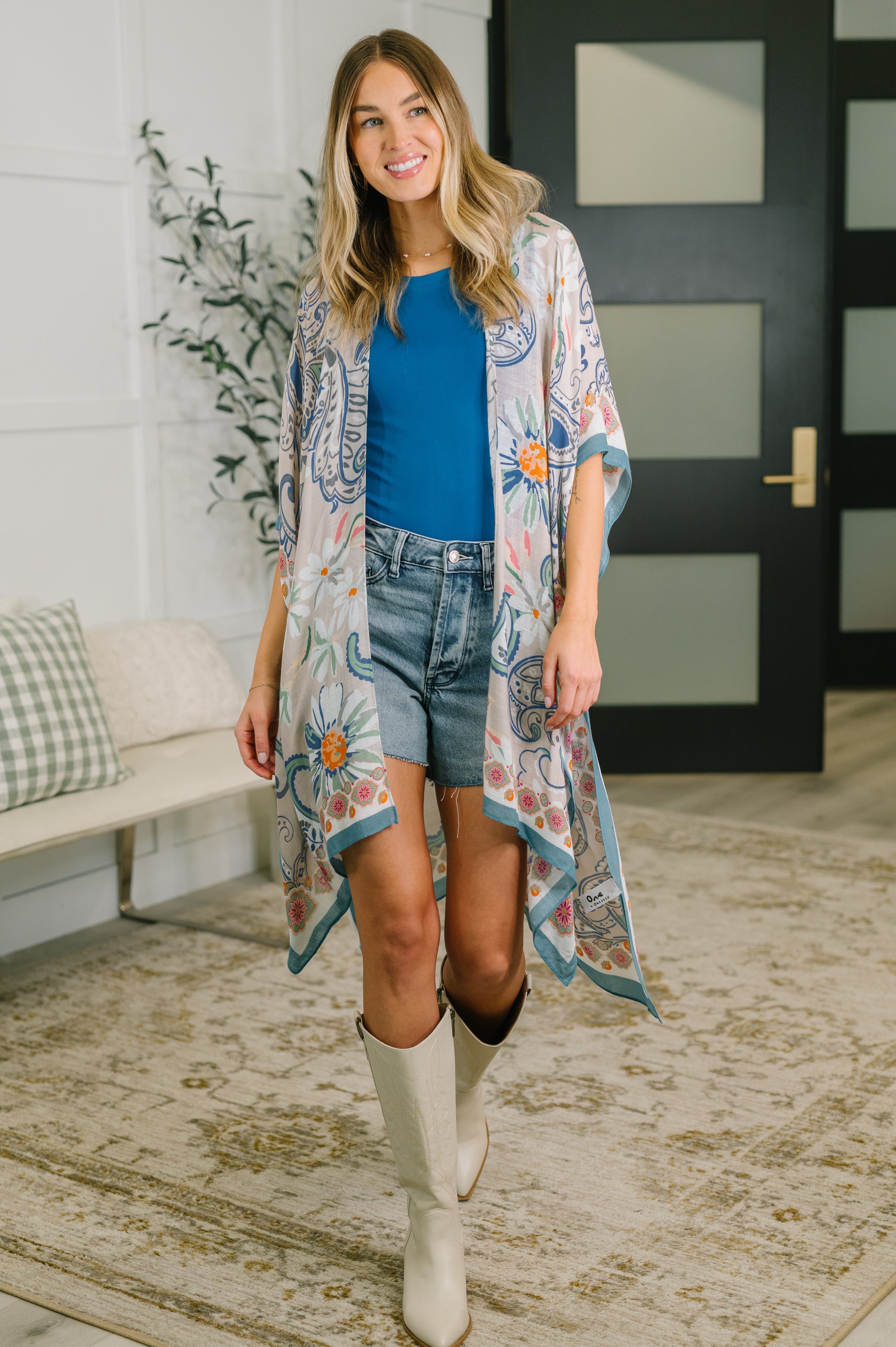 Woman wearing blue layering piece with floral print over blue top and denim shorts in cozy indoor setting