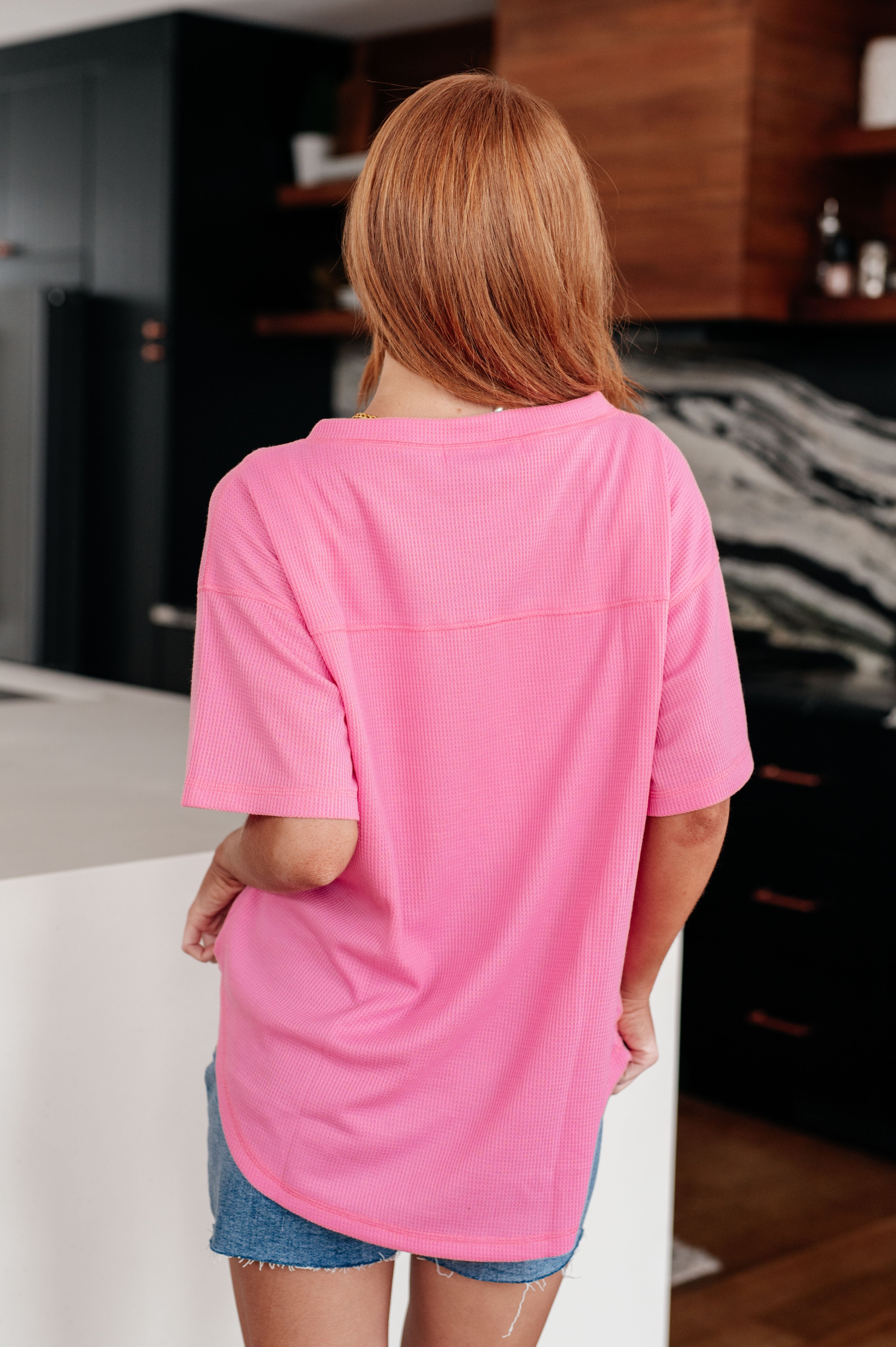 Woman with long red hair wearing bright pink Back Together V-Neck Top, back view, standing indoors in modern kitchen setting