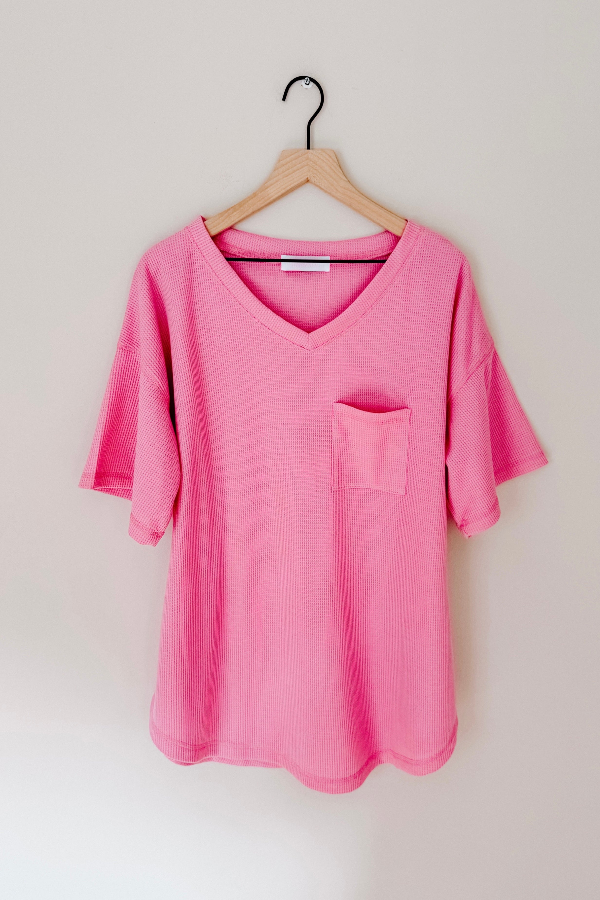 Pink Back Together V-Neck Top with front pocket hanging on wooden hanger against beige wall, gallery image 0