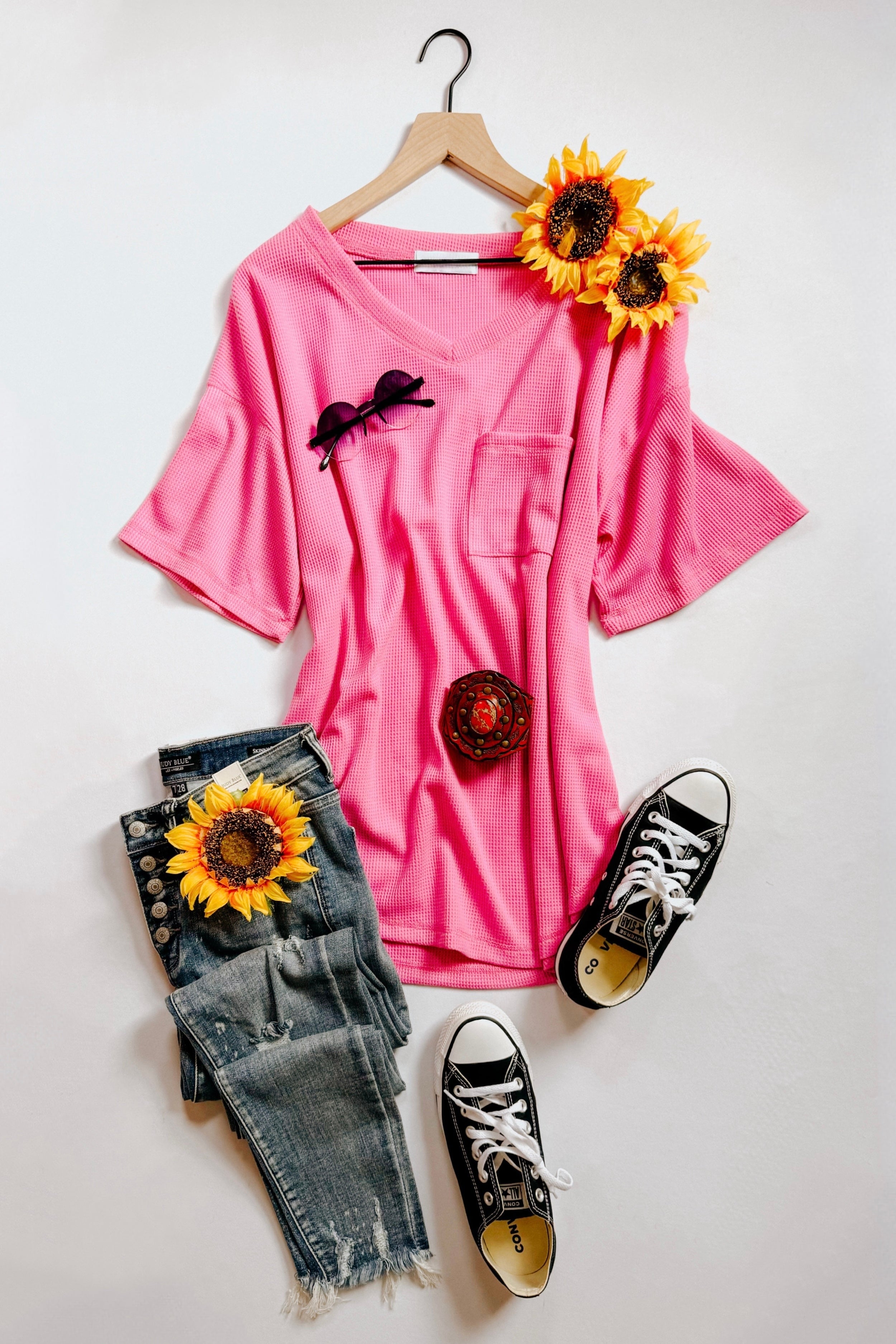 Bright pink Back Together V-Neck Top displayed on hanger with sunglasses, denim jeans, sneakers, and sunflowers flat lay