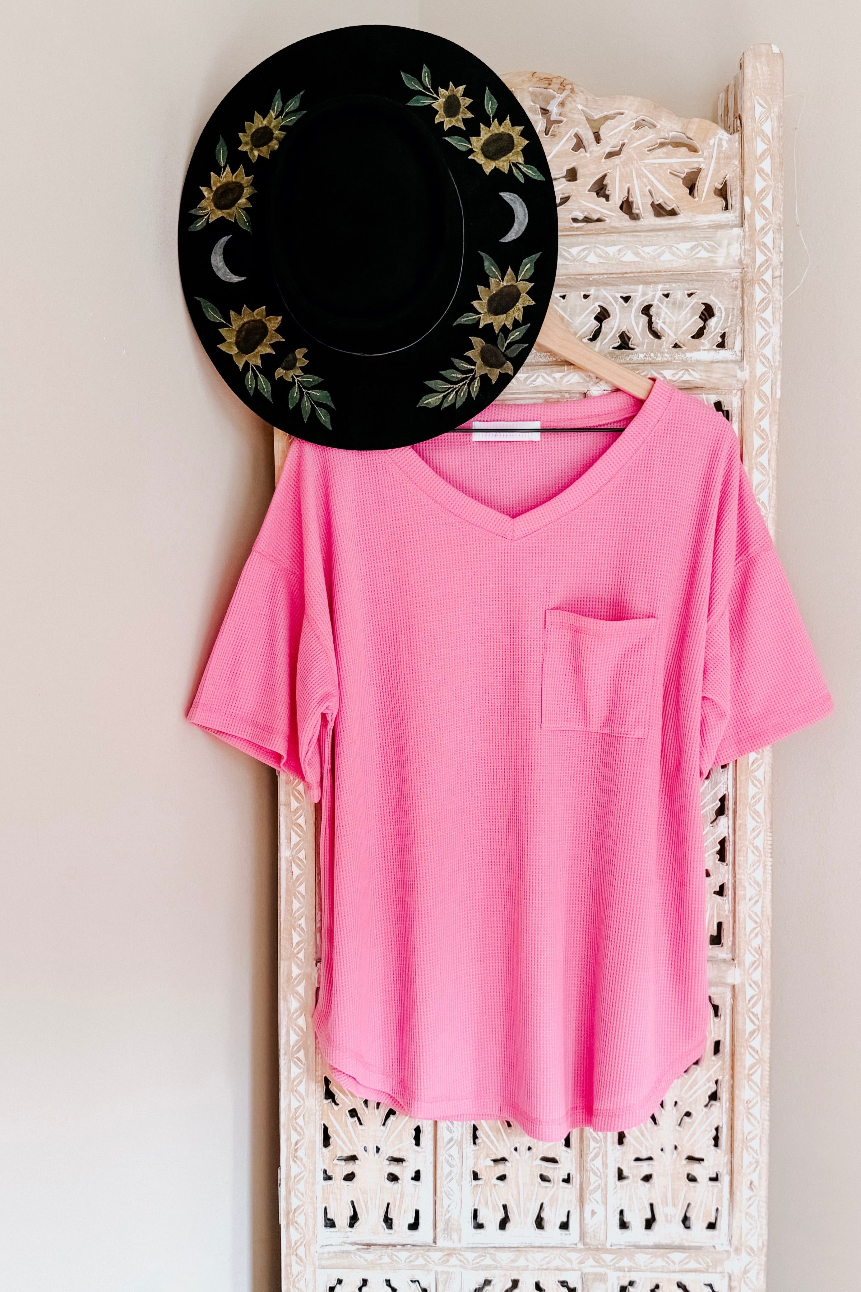 Bright pink Back Together V-Neck Top with front pocket hanging on carved white wooden panel with black floral hat above