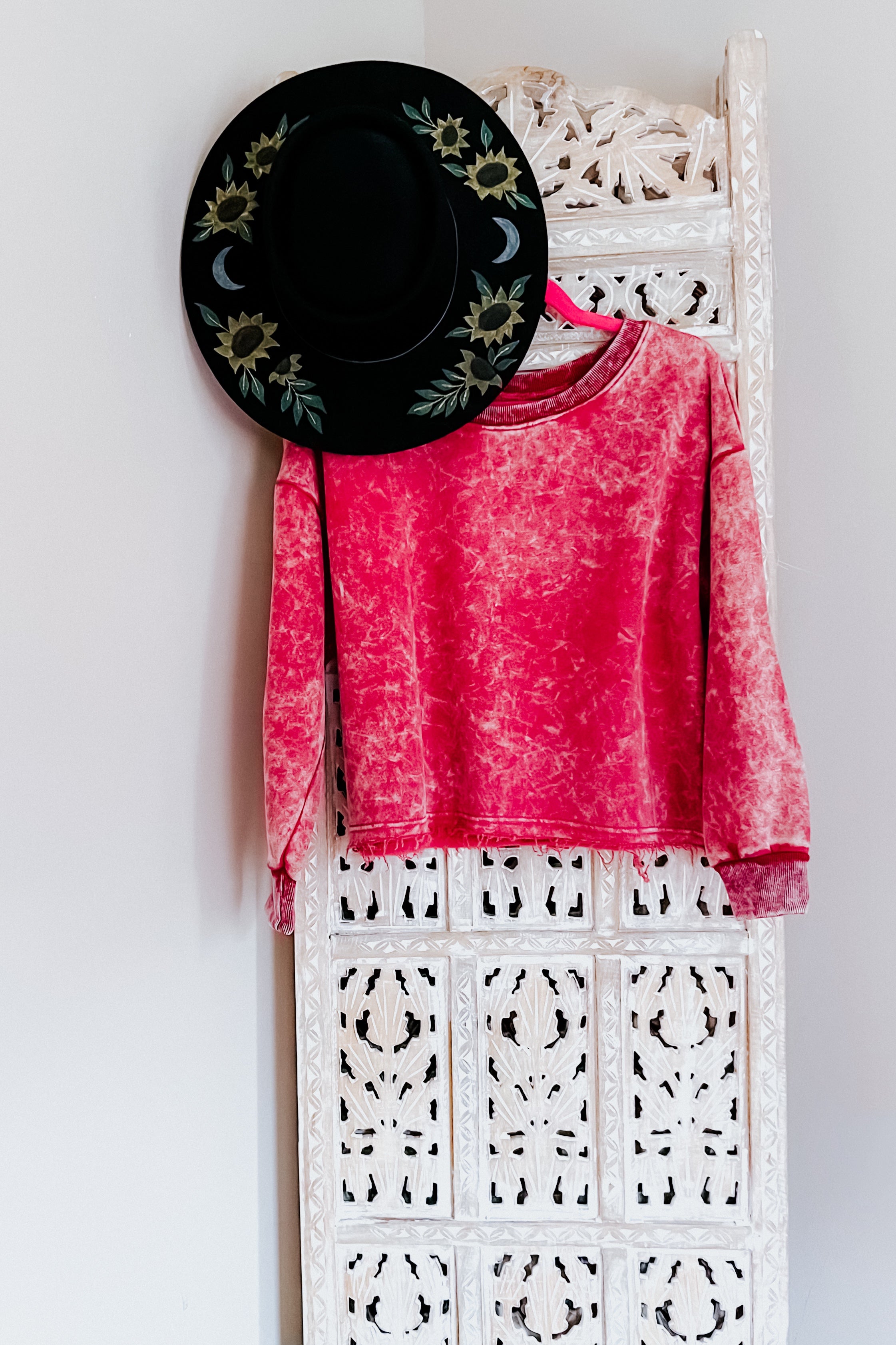 Berry acid wash cropped pullover hanging on ornate white wooden panel with black floral and moon embroidered hat above