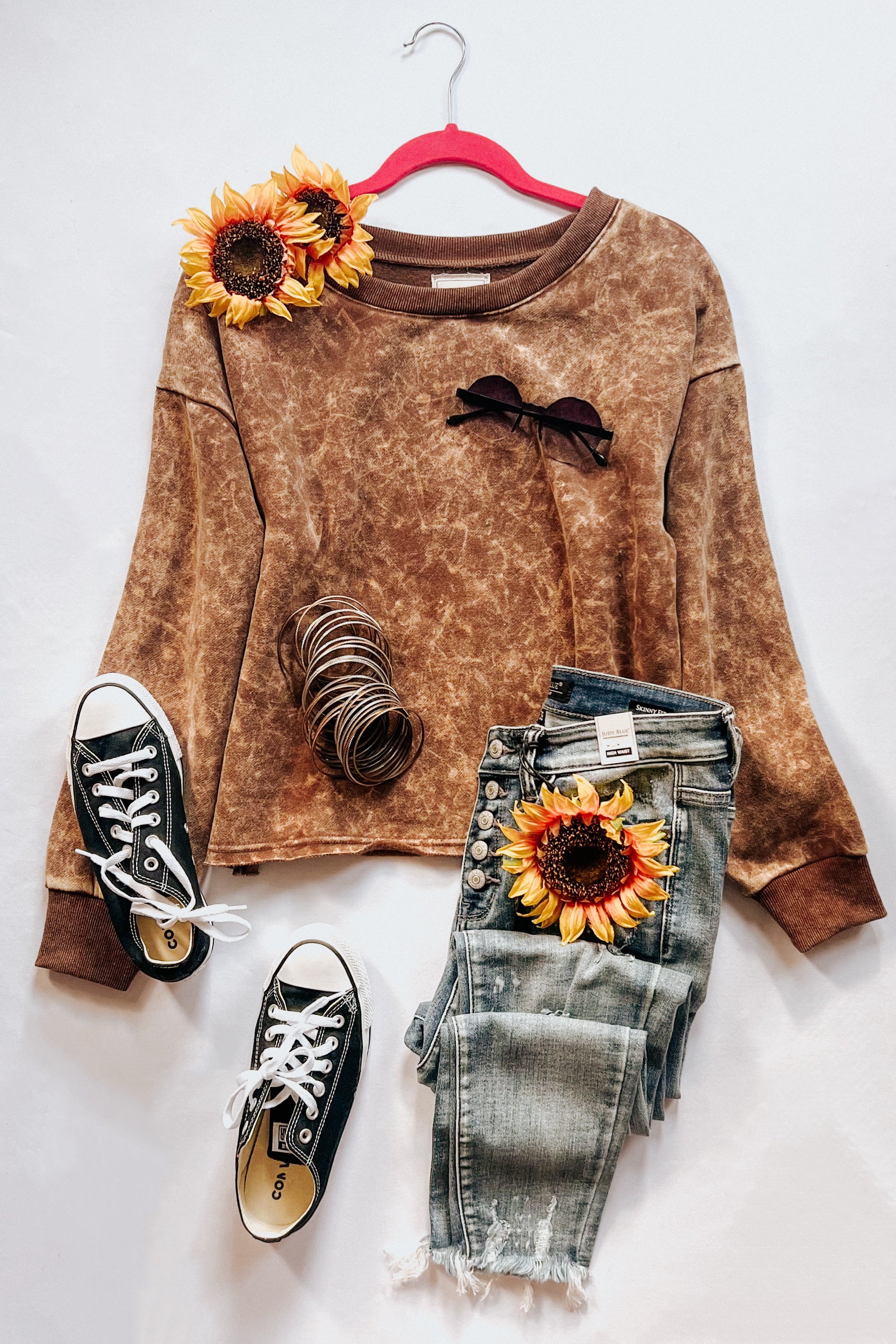 Mocha acid wash pullover on pink hanger styled with distressed jeans, black sneakers, bracelets, sunglasses, and sunflowers