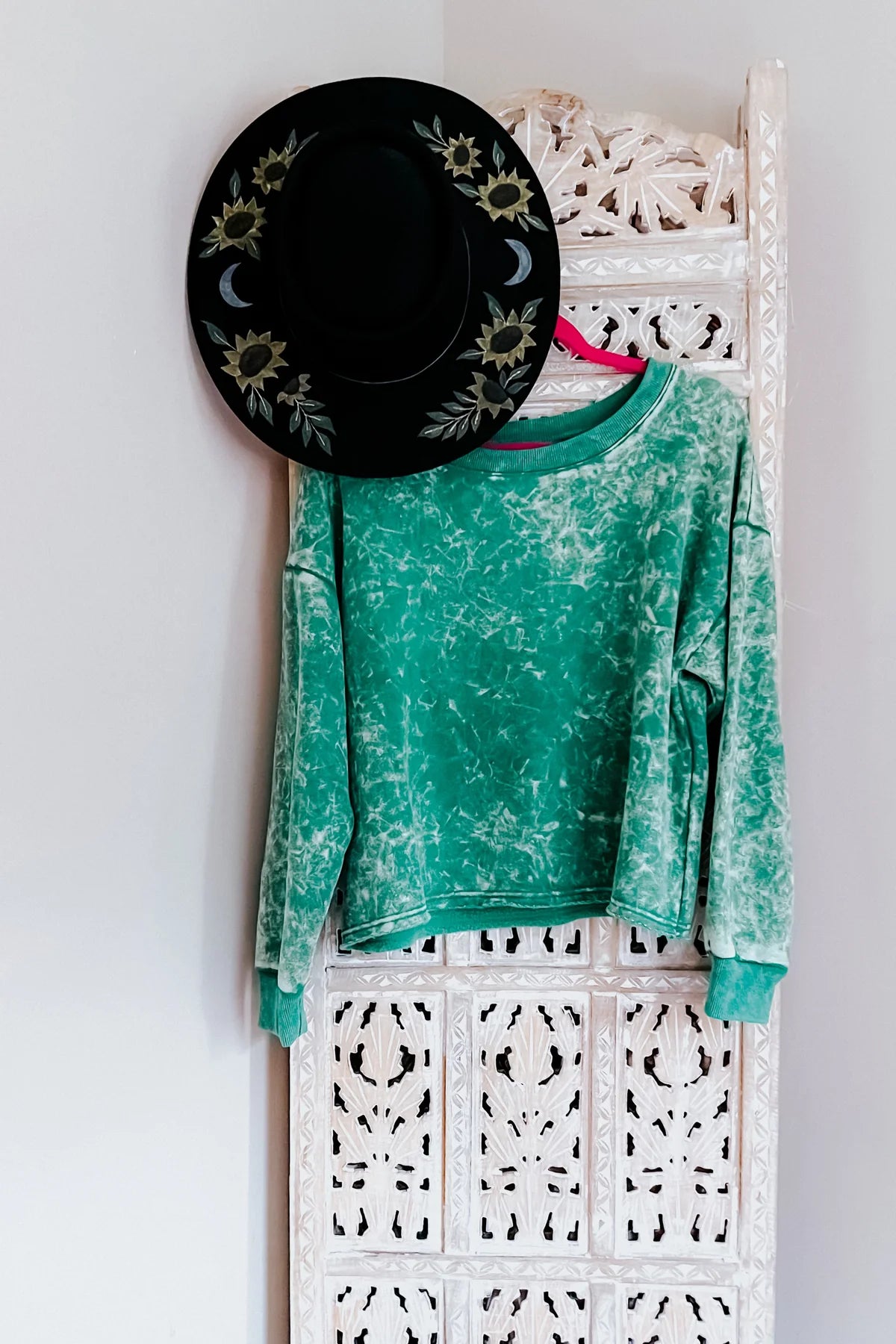 Sea green acid wash cropped pullover hanging on carved white wooden panel with black hat featuring sunflowers and crescent moons