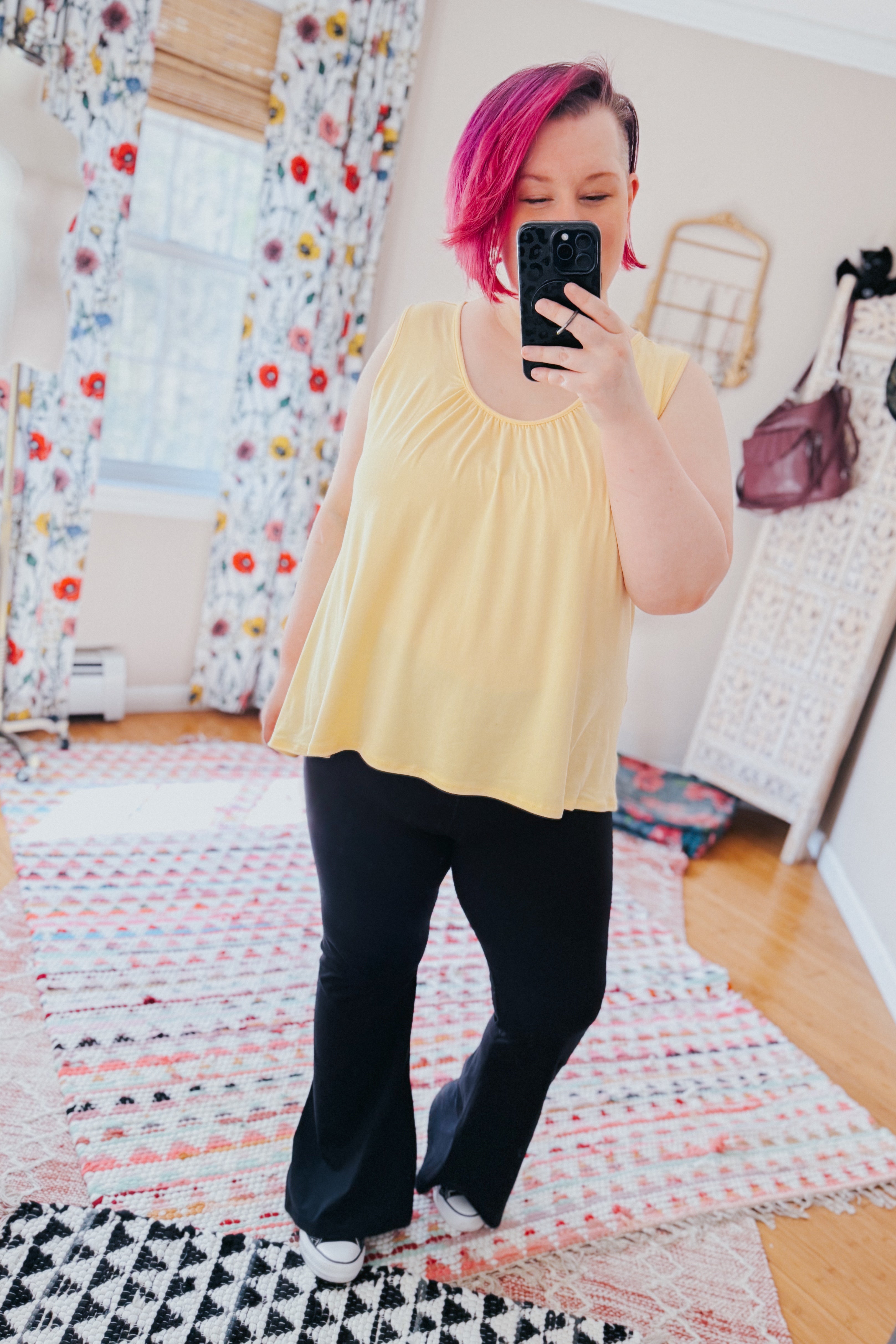 Person with short pink hair wearing loose soft yellow Banana Bliss tank top and black pants taking indoor mirror selfie.