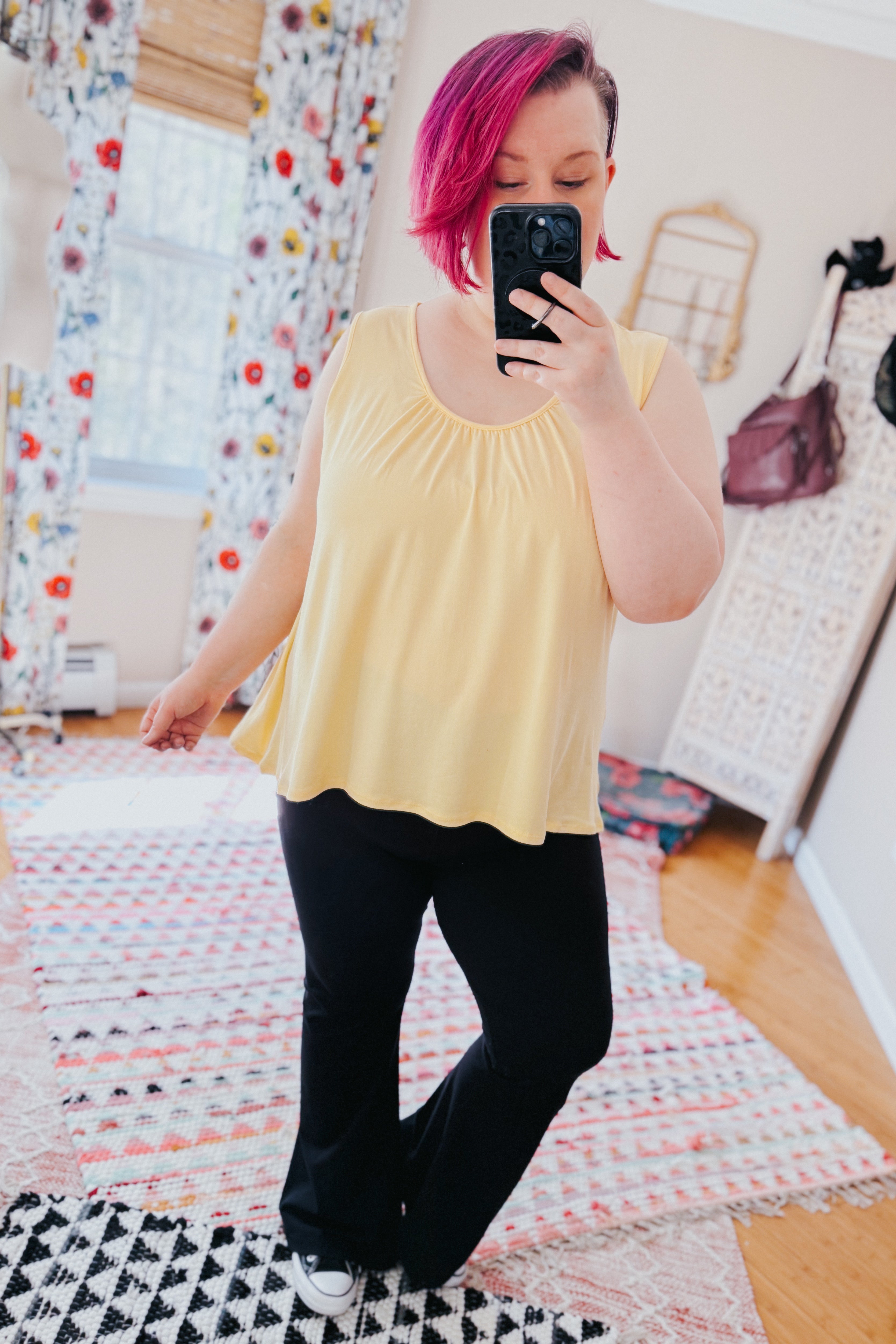 Person with pink hair taking indoor mirror selfie wearing loose yellow Banana Bliss tank top and black pants