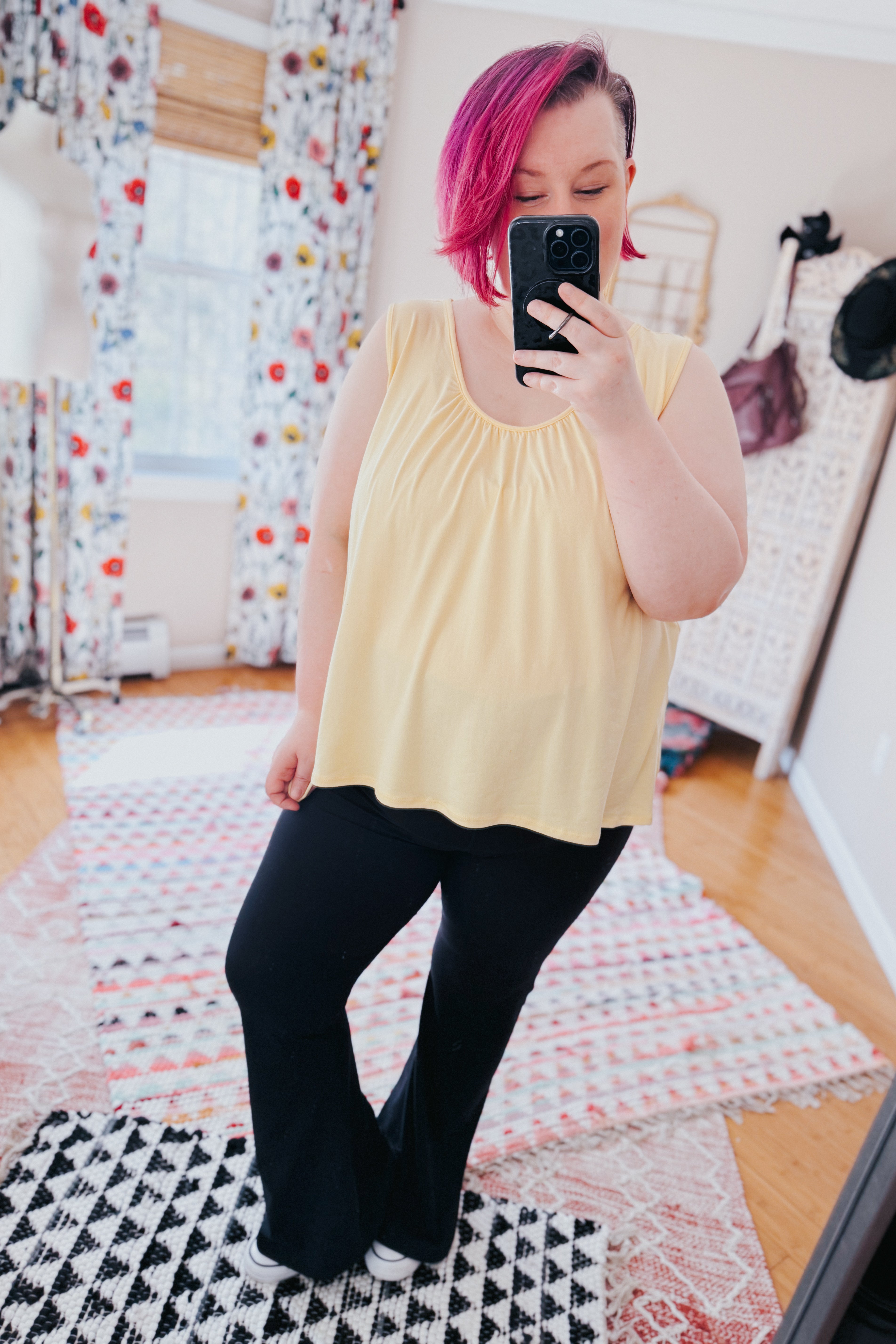 Person with bright pink hair taking mirror selfie wearing loose yellow Banana Bliss tank top and black pants indoors