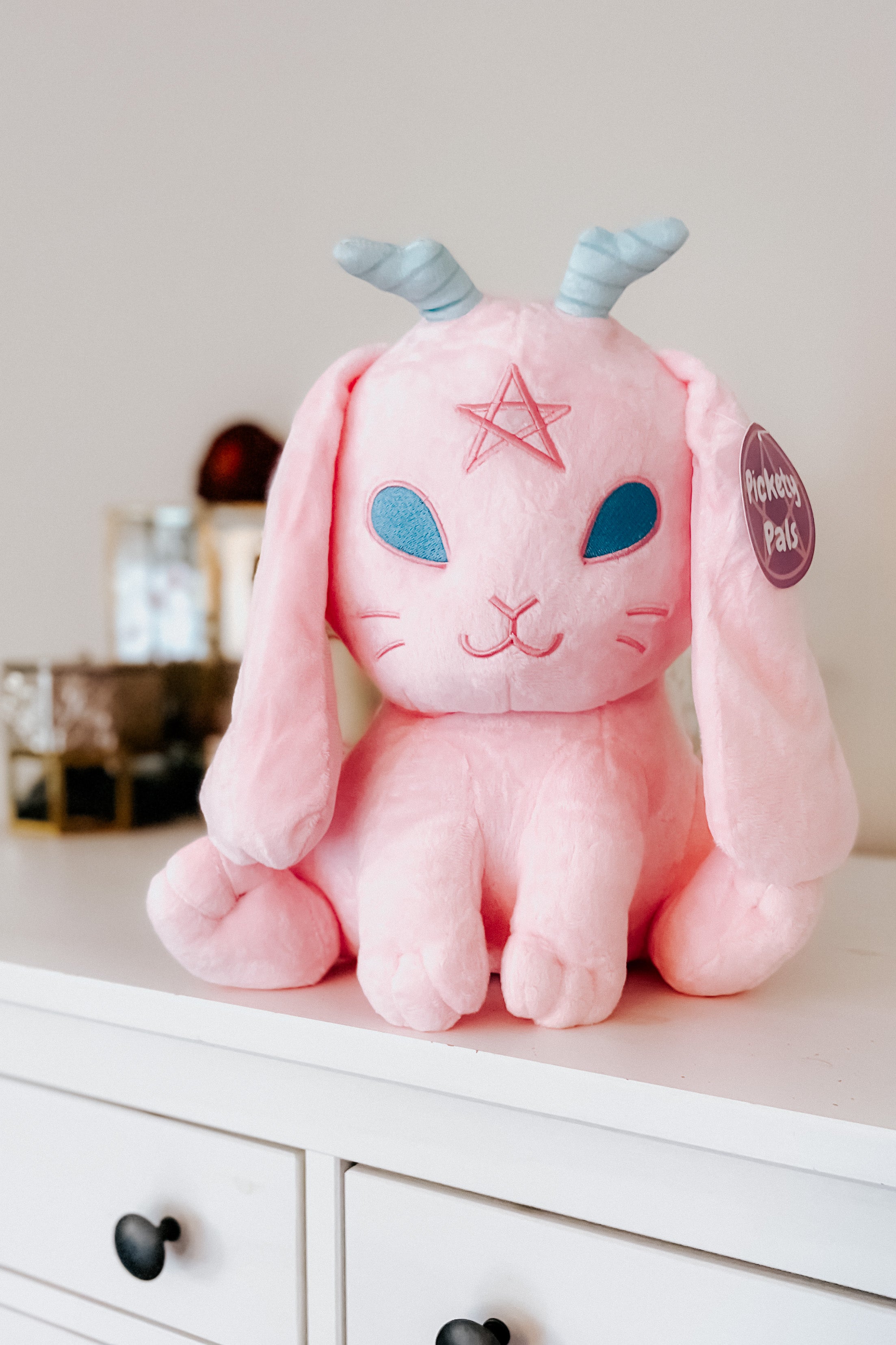 Front angle of cotton candy pink Beelzebun Witchy Bunny plush with blue eyes and spiral horns on white dresser surface