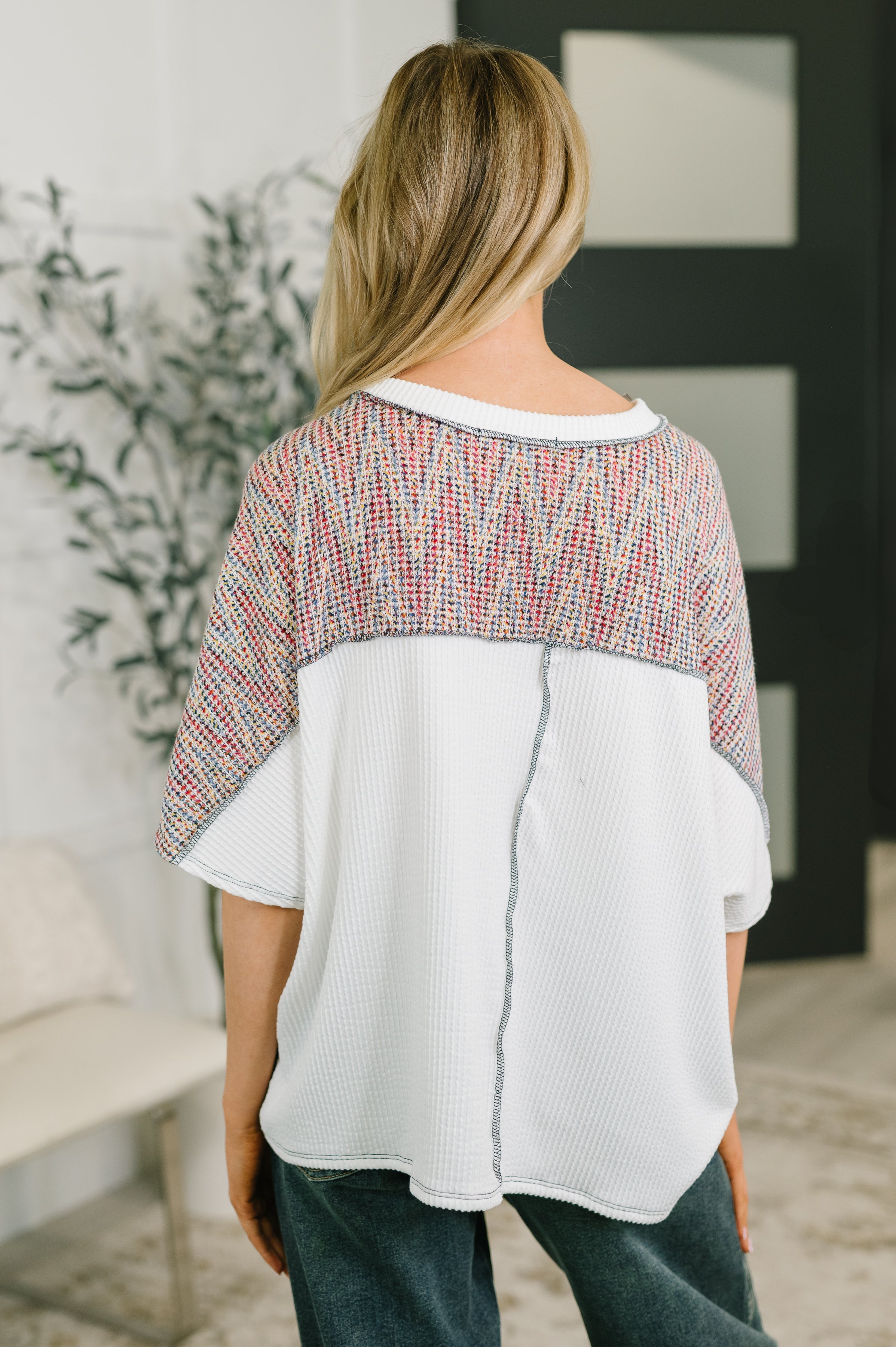 Back view of woman wearing loose white textured top with colorful knit yoke and dolman sleeves indoors