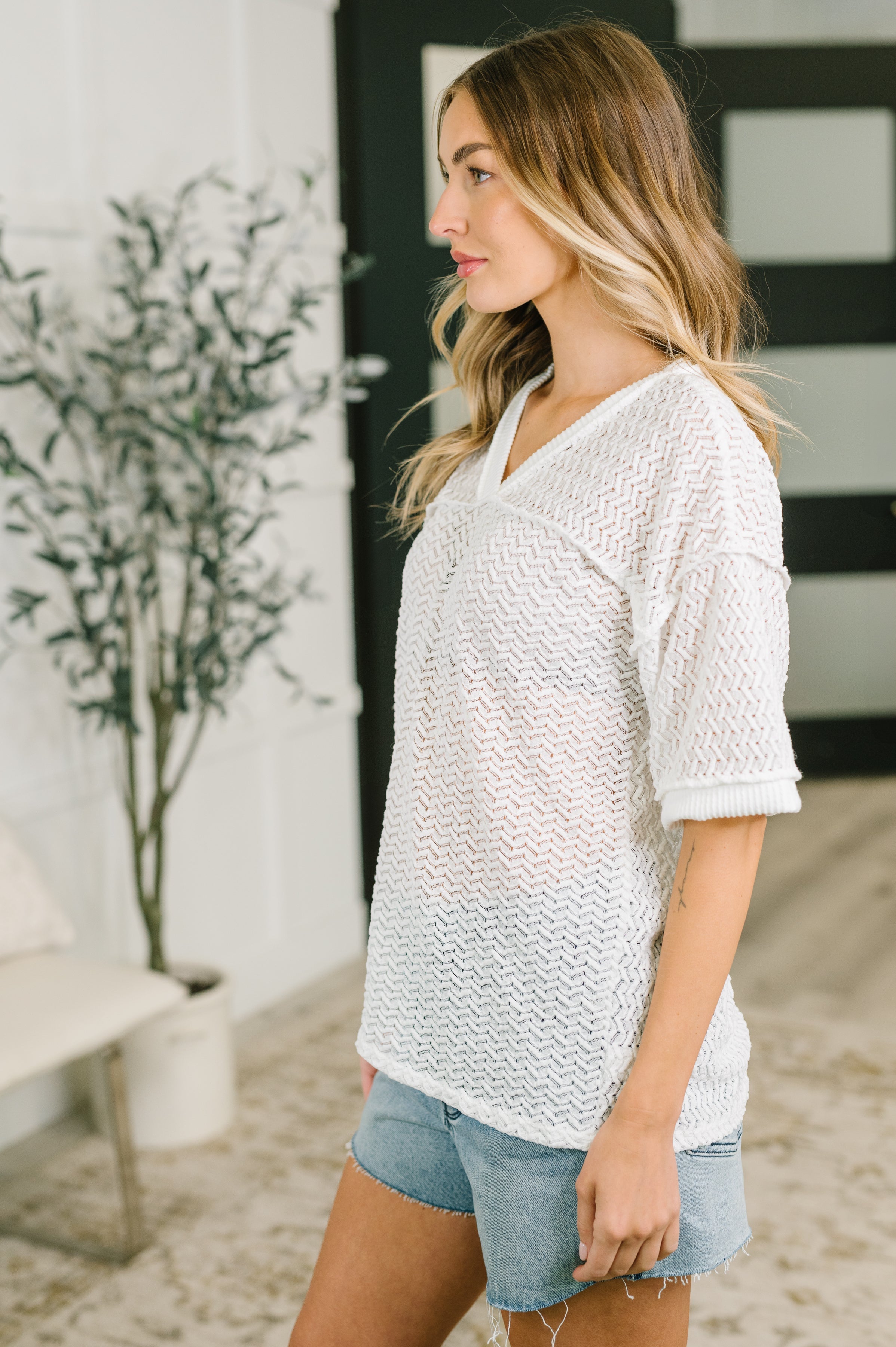 Woman wearing cream textured oversized top with V-neck and rolled sleeves paired with denim shorts indoors