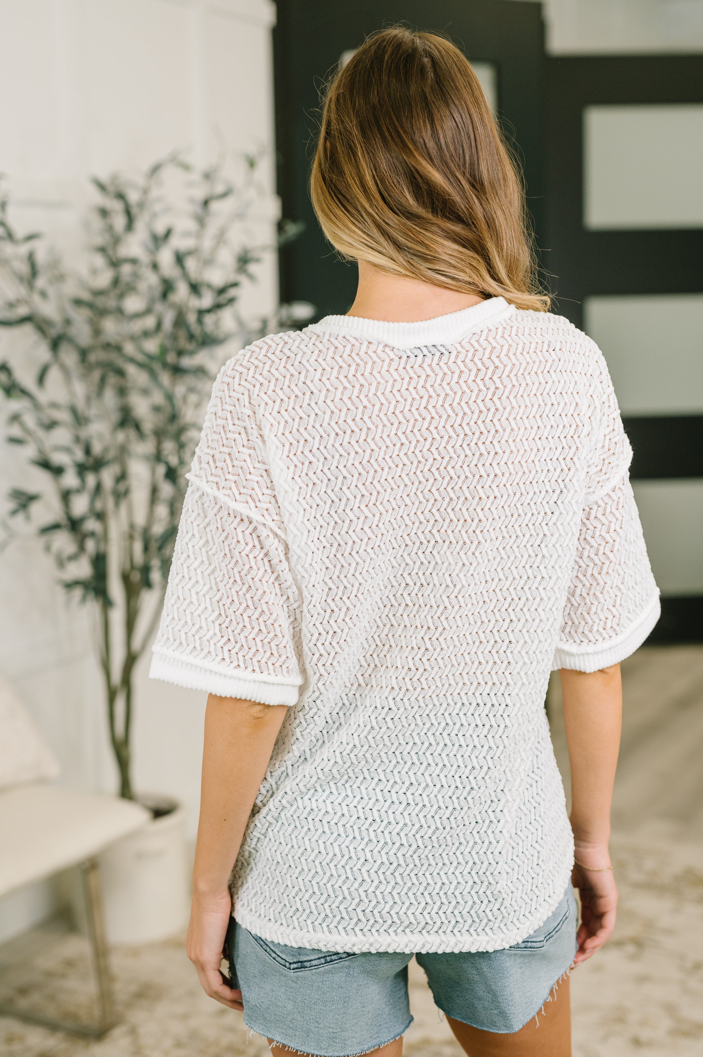 Back view of woman wearing cream textured oversized top with short sleeves and cuffed edges paired with denim shorts indoors