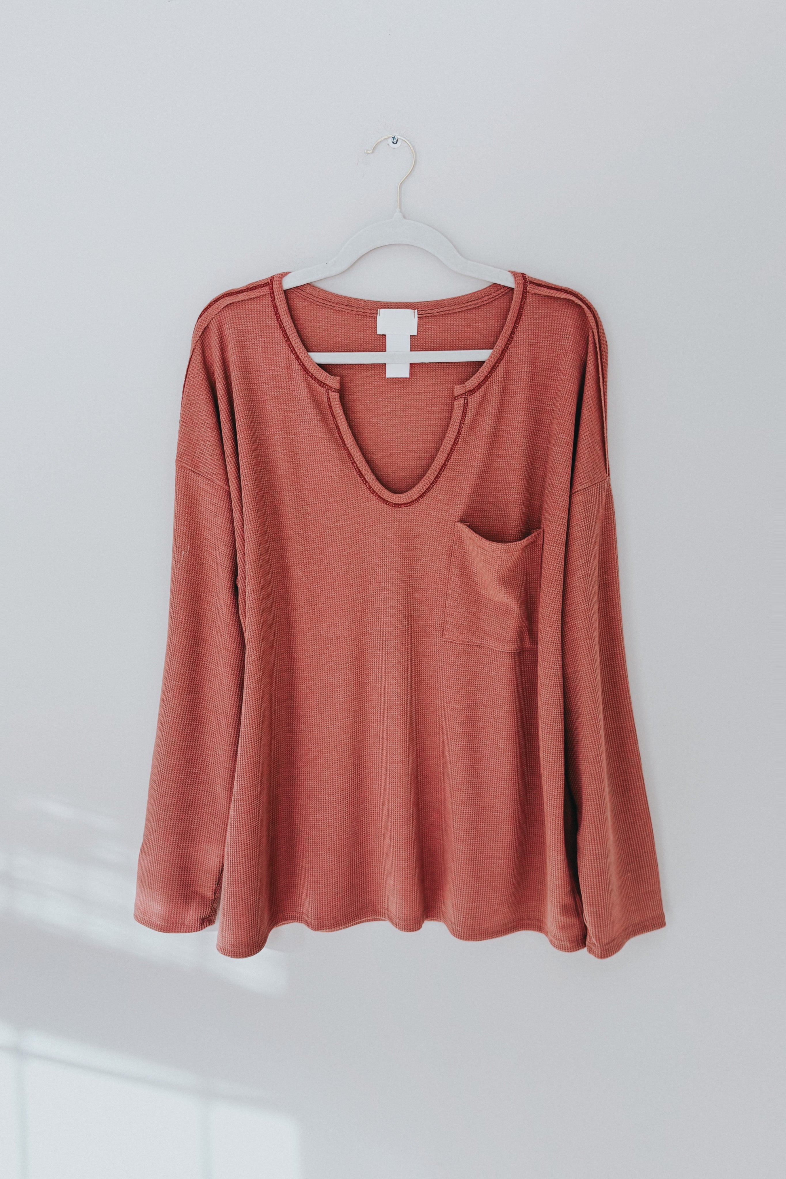 Front view of rust-colored waffle knit top with pocket hanging on white hanger against gray wall