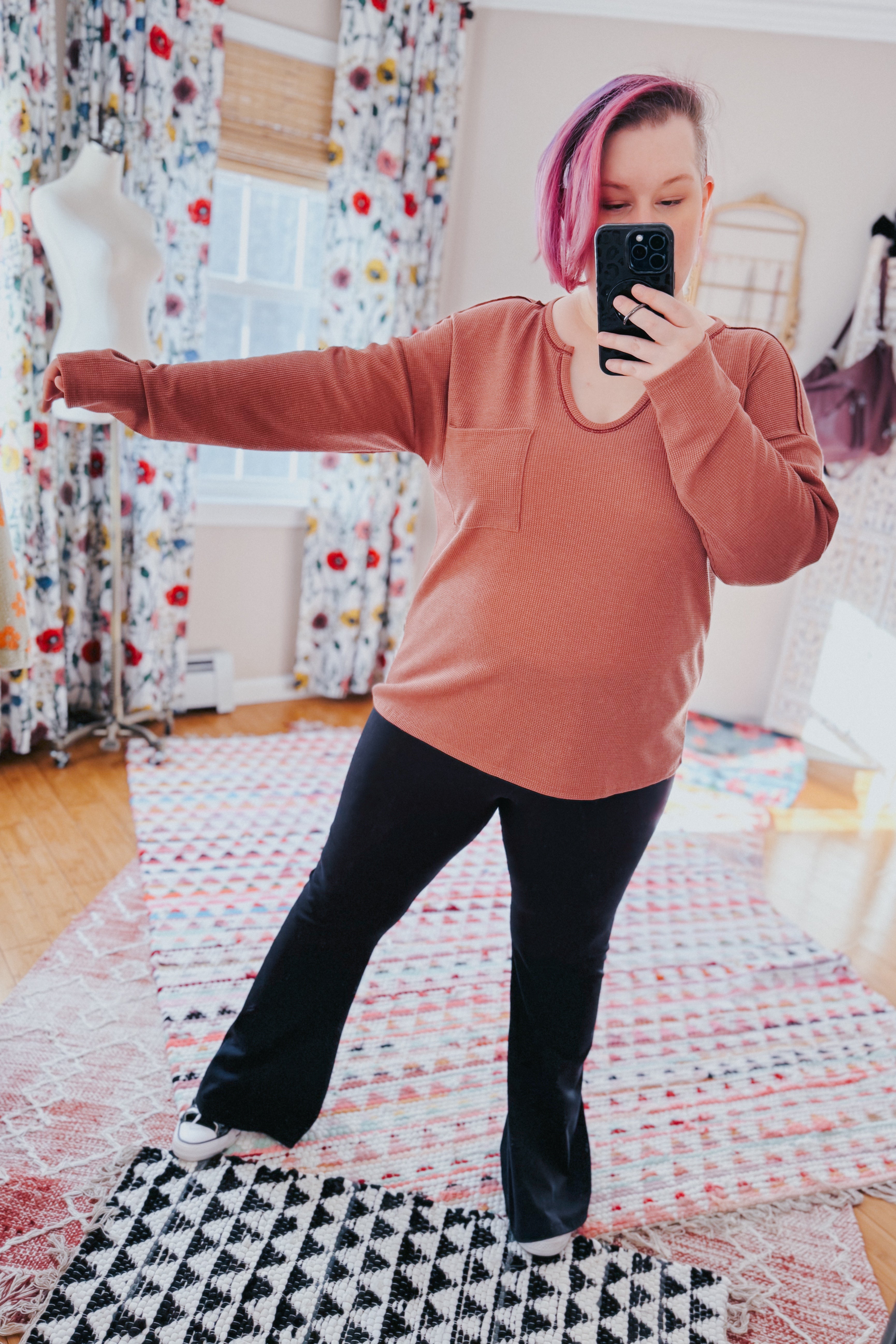 Woman wearing rust-colored Behind The Scenes waffle knit top with pocket taking selfie in bright room with floral curtains