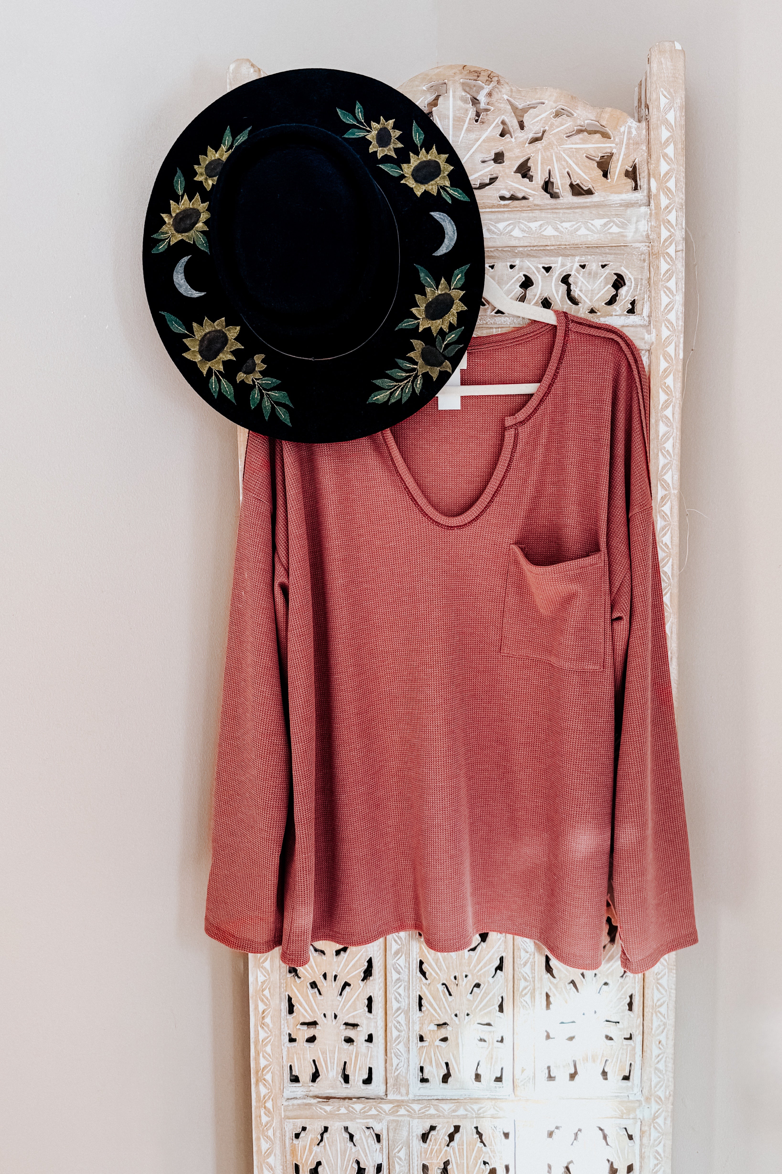Rust-colored waffle knit top with pocket hanging on carved white wooden screen with black hat decorated with sunflowers