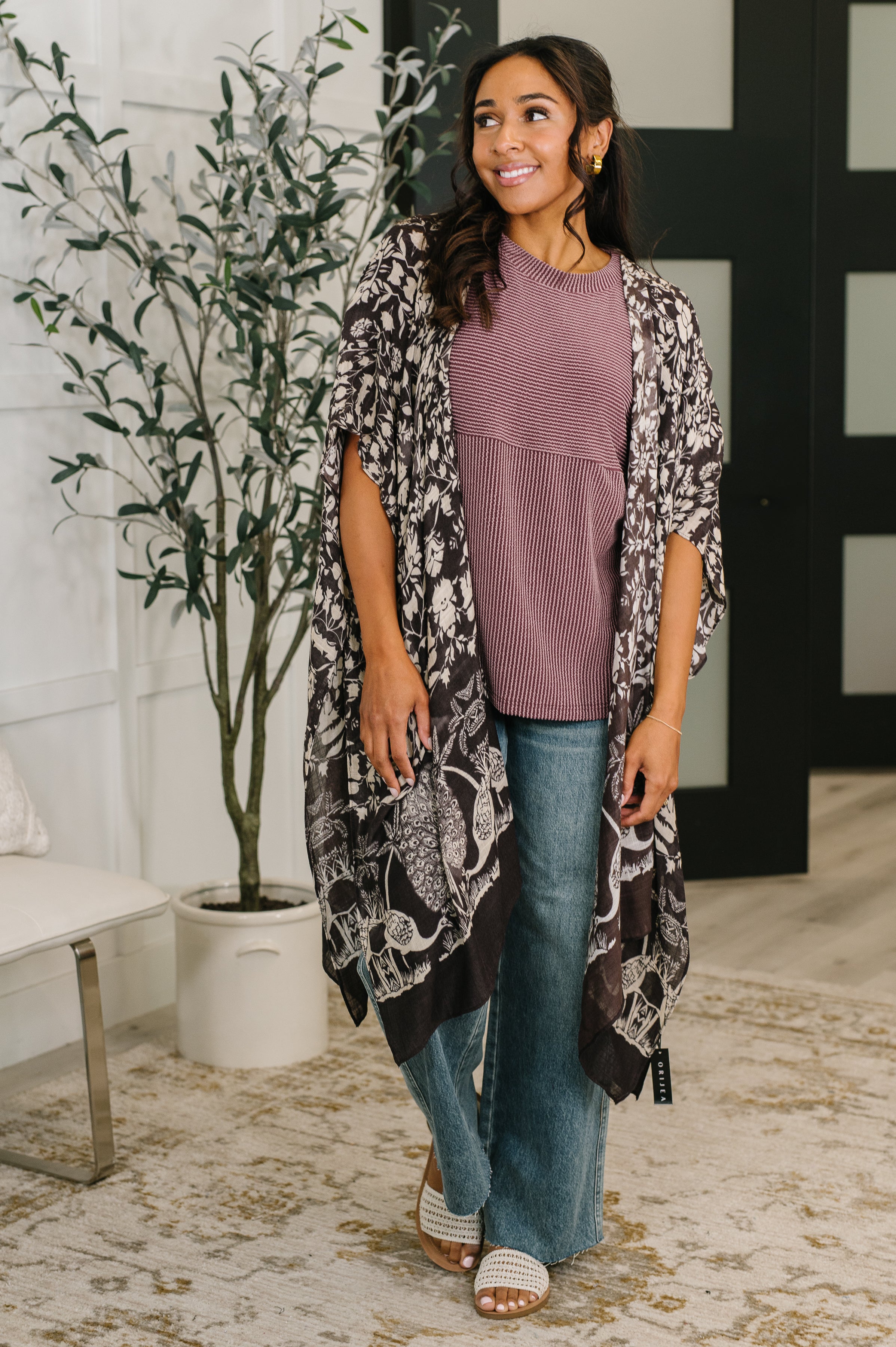 Woman wearing black floral open front duster over mauve top and wide-leg jeans in indoor setting with plant and rug