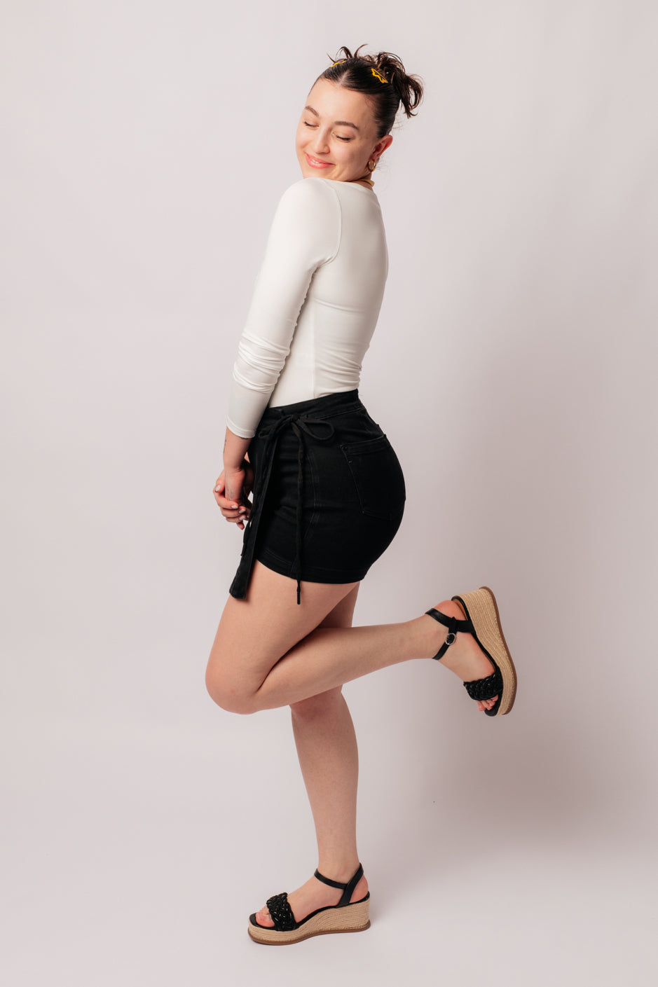 Model posing in Bentli black high rise denim skort with tied side detail paired with white long sleeve top and black wedge sandals