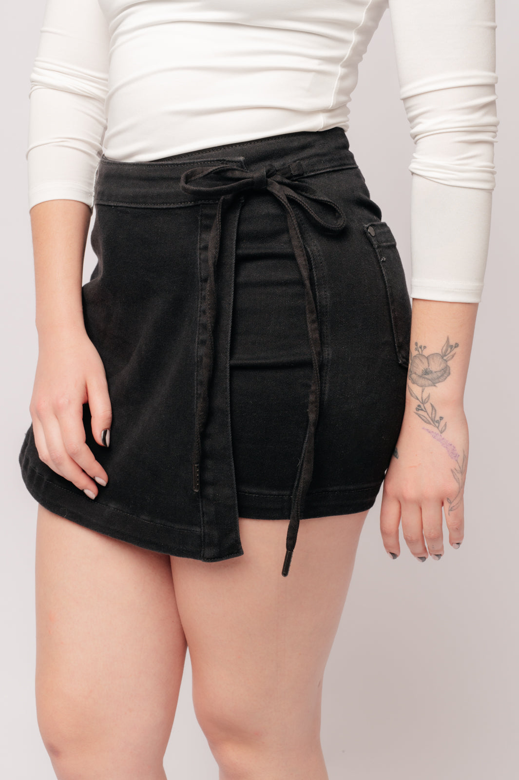 Woman wearing Bentli high rise black denim skort with side tie detail and floral tattoo on arm against neutral background