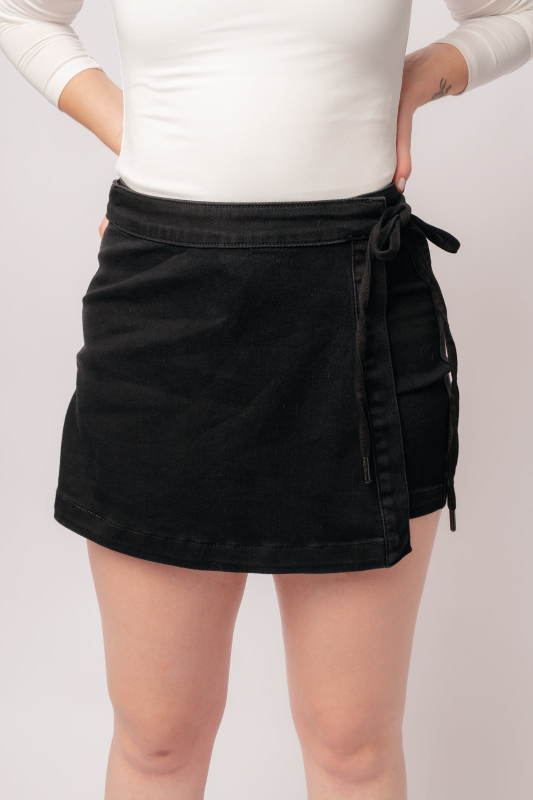 Front view of Bentli High Rise black denim skort with side tie detail worn with white top against neutral background