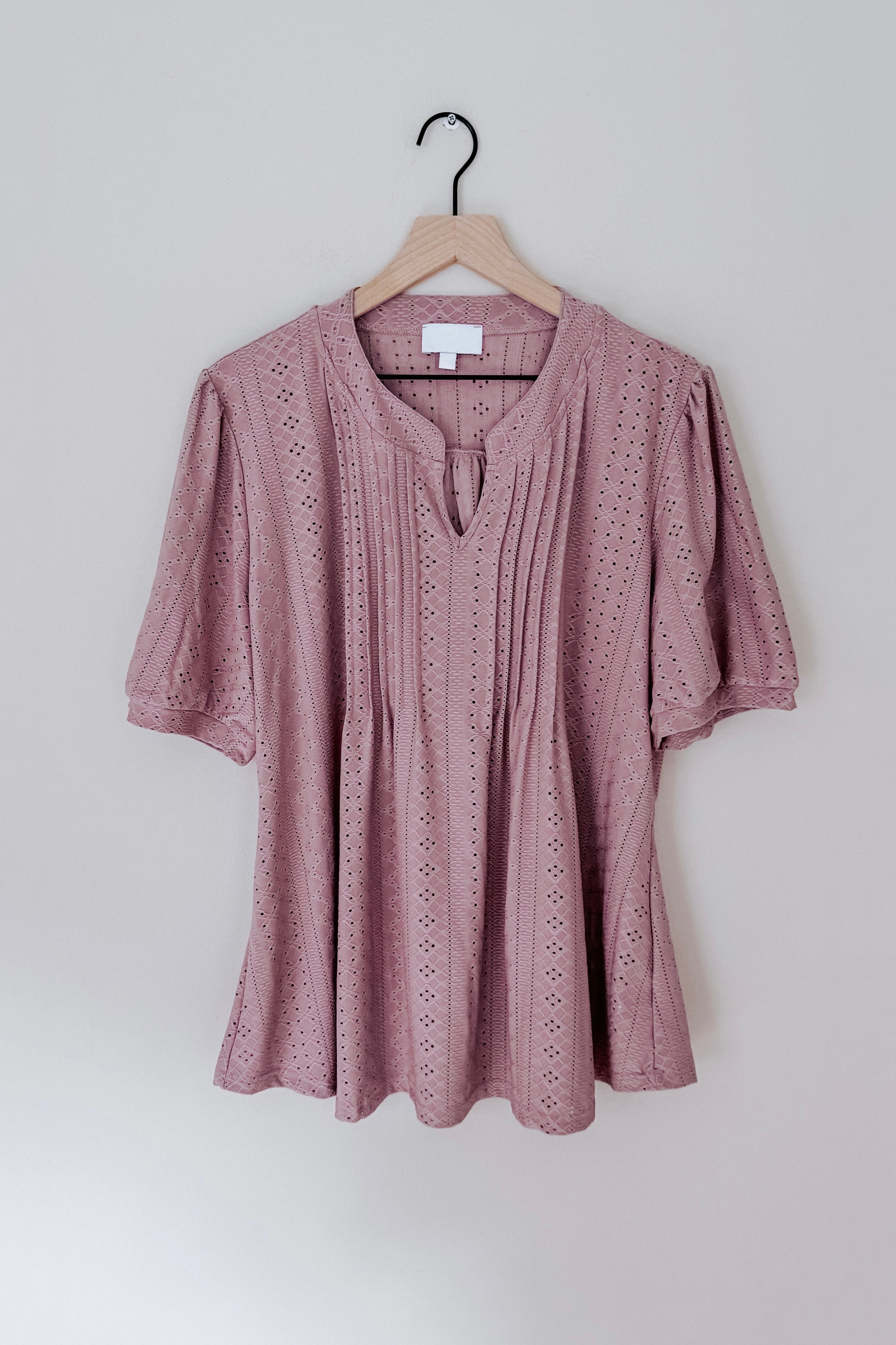 Mauve pleated front blouse with eyelet pattern and short puff sleeves hanging on wooden hanger against pale wall