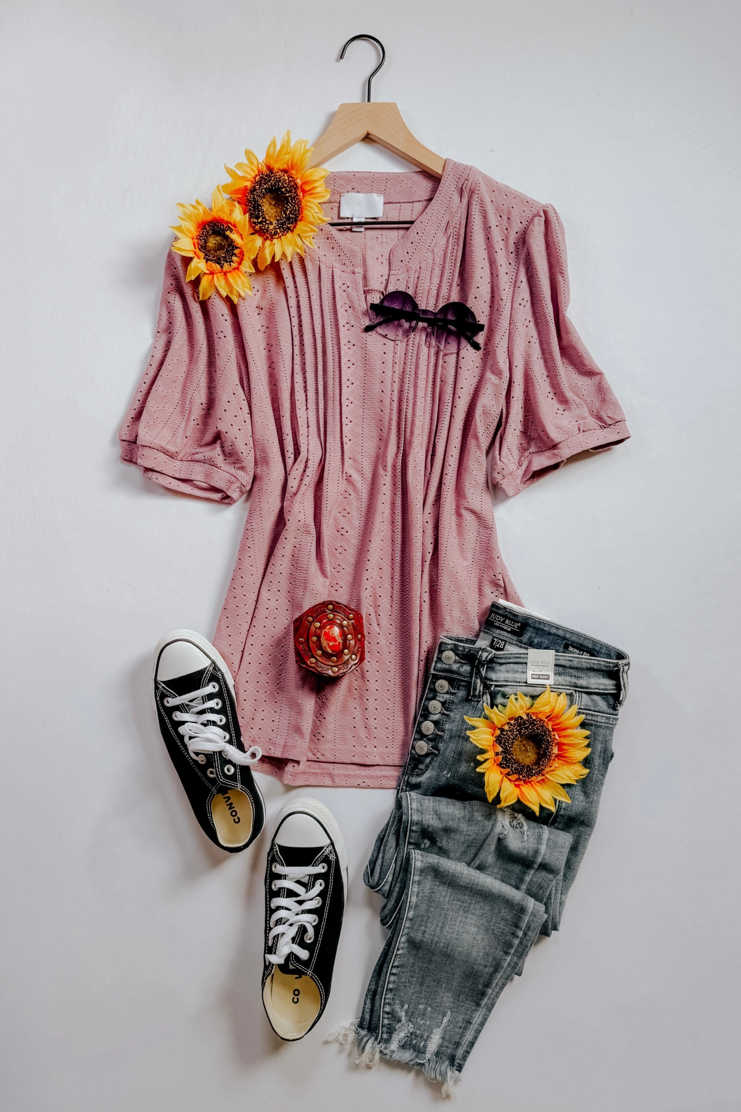 Pink pleated front blouse with eyelet texture styled with distressed jeans, black sneakers, sunglasses, and sunflower accents