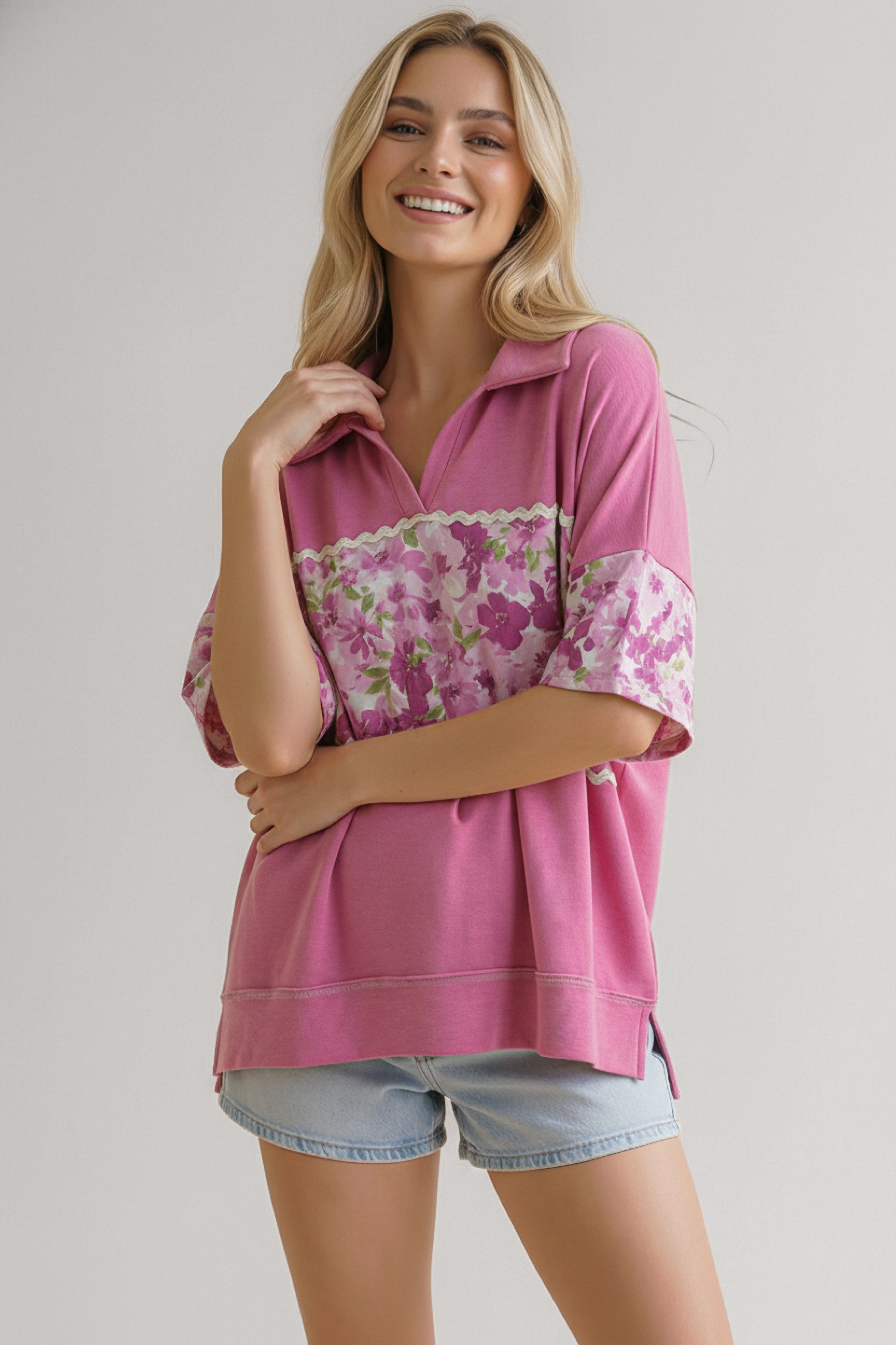 Smiling woman wearing a pink top with floral band and collar paired with light denim shorts standing against plain background