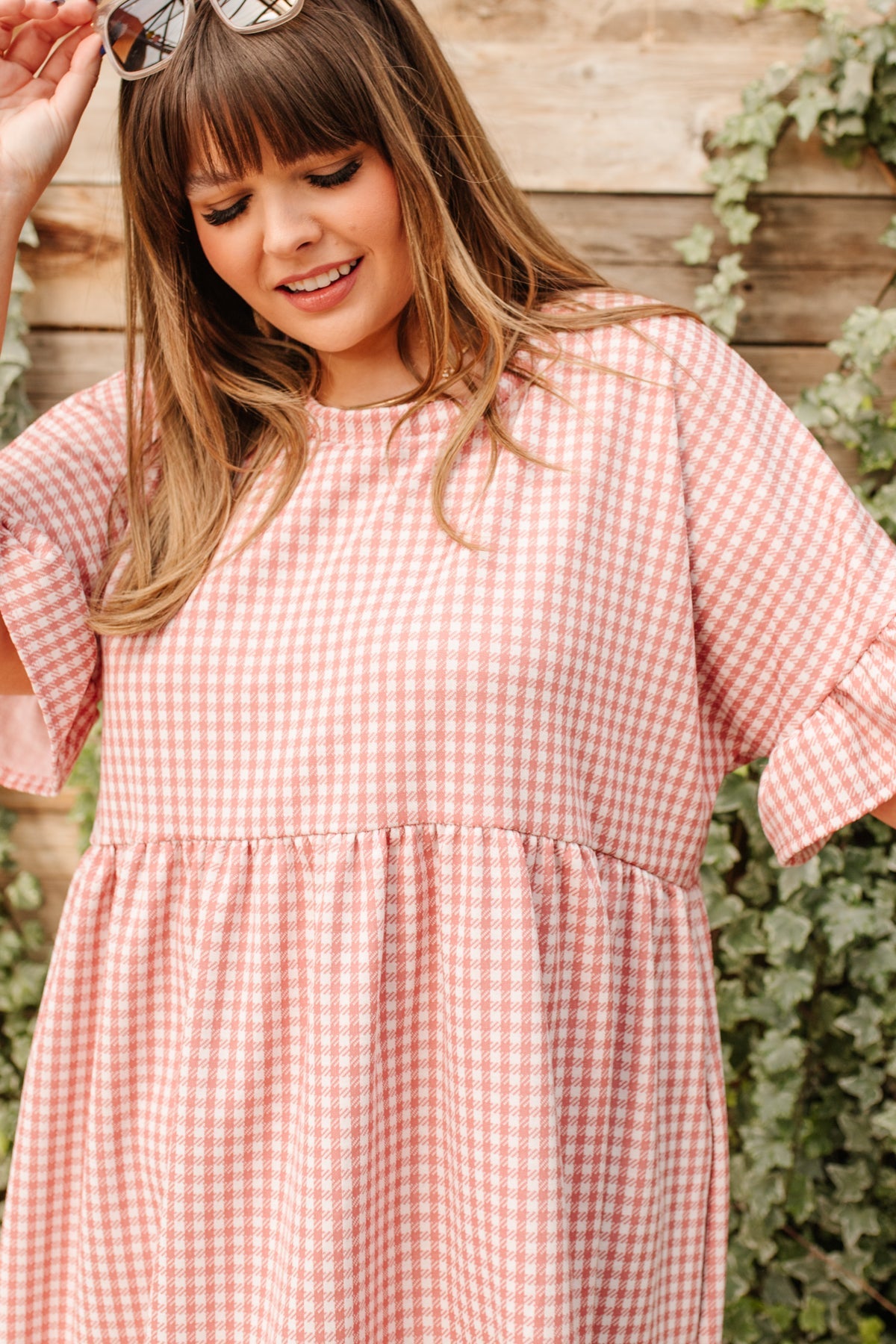 Smiling woman wearing pink and white gingham dress with loose ruffle sleeves standing near ivy and wooden fence