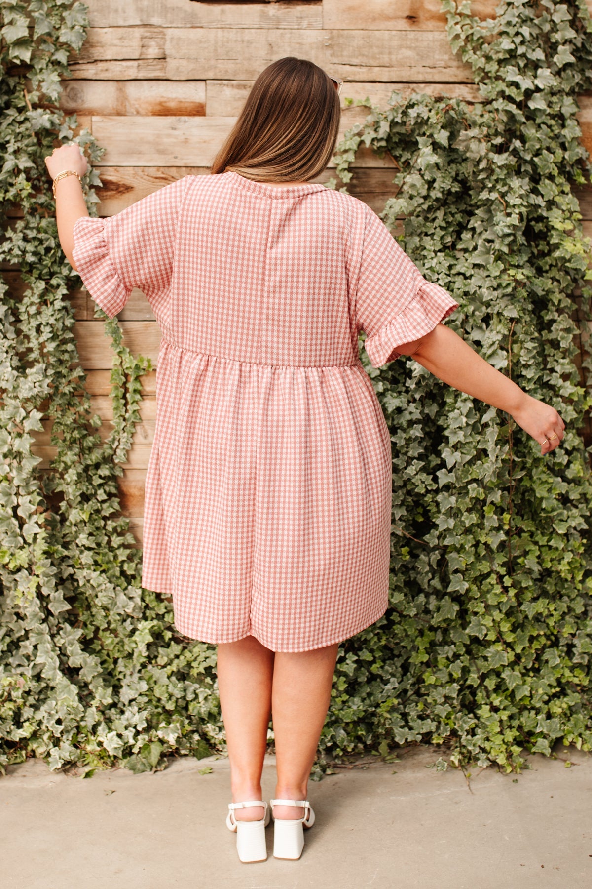 Woman wearing pink gingham short sleeve dress with ruffle detail and white block heel shoes standing outdoors