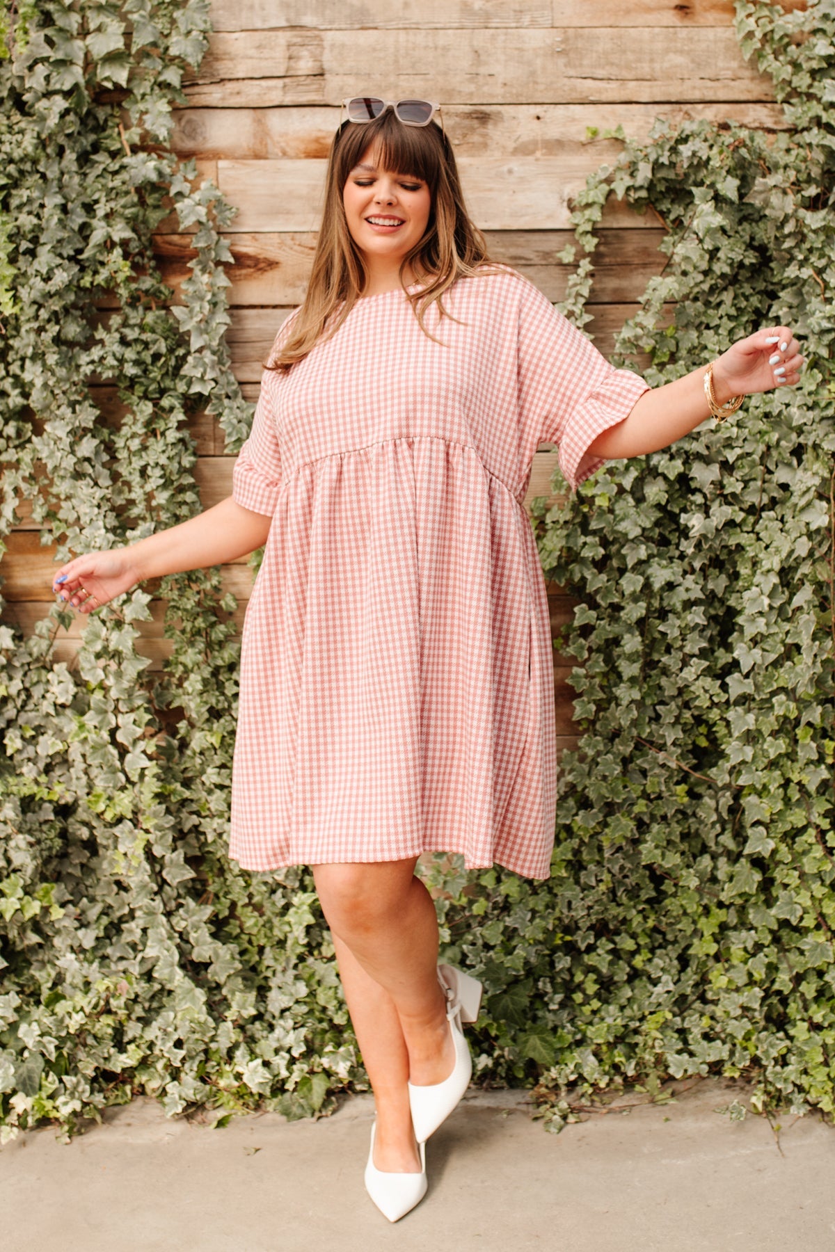 Woman wearing loose pink gingham dress with elbow-length sleeves and white pointed toe heels by ivy-covered wooden wall