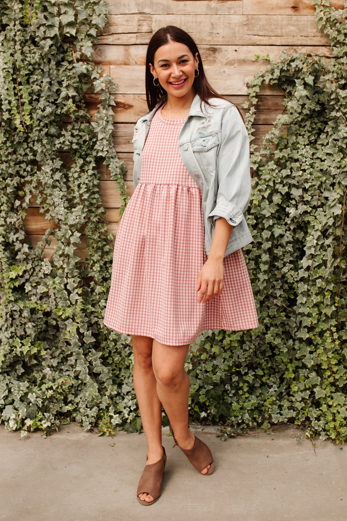 Smiling woman in short pink gingham dress with a light denim jacket and brown peep-toe booties by ivy-covered wood wall
