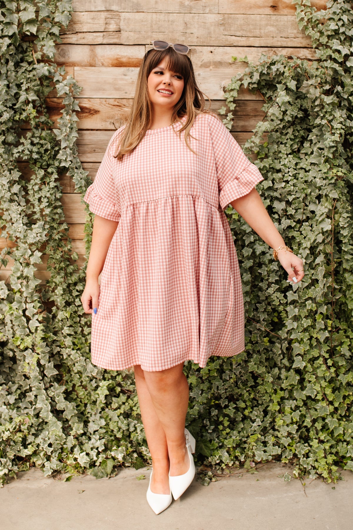 Plus size woman wearing loose pink gingham dress with ruffle sleeves and white pointed toe heels next to ivy-covered wood wall