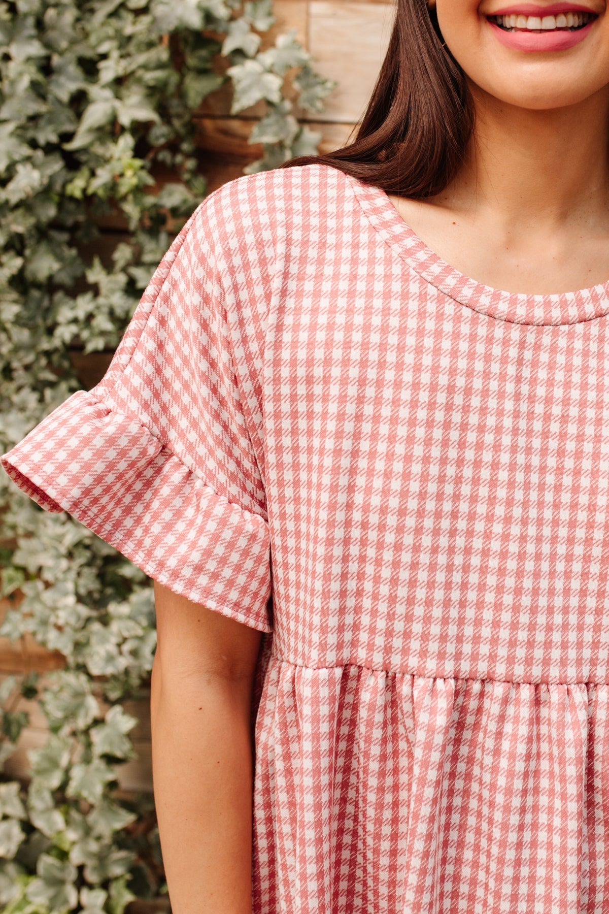 Woman wearing pink and white checkered dress with short cuffed sleeves standing by wooden wall with ivy leaves