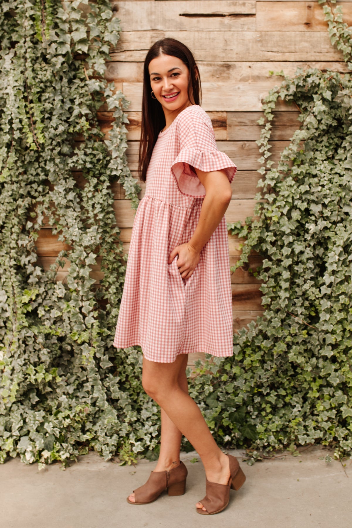 Woman wearing pink gingham babydoll dress with ruffle sleeves and brown open-toe block heels standing by ivy-covered wall