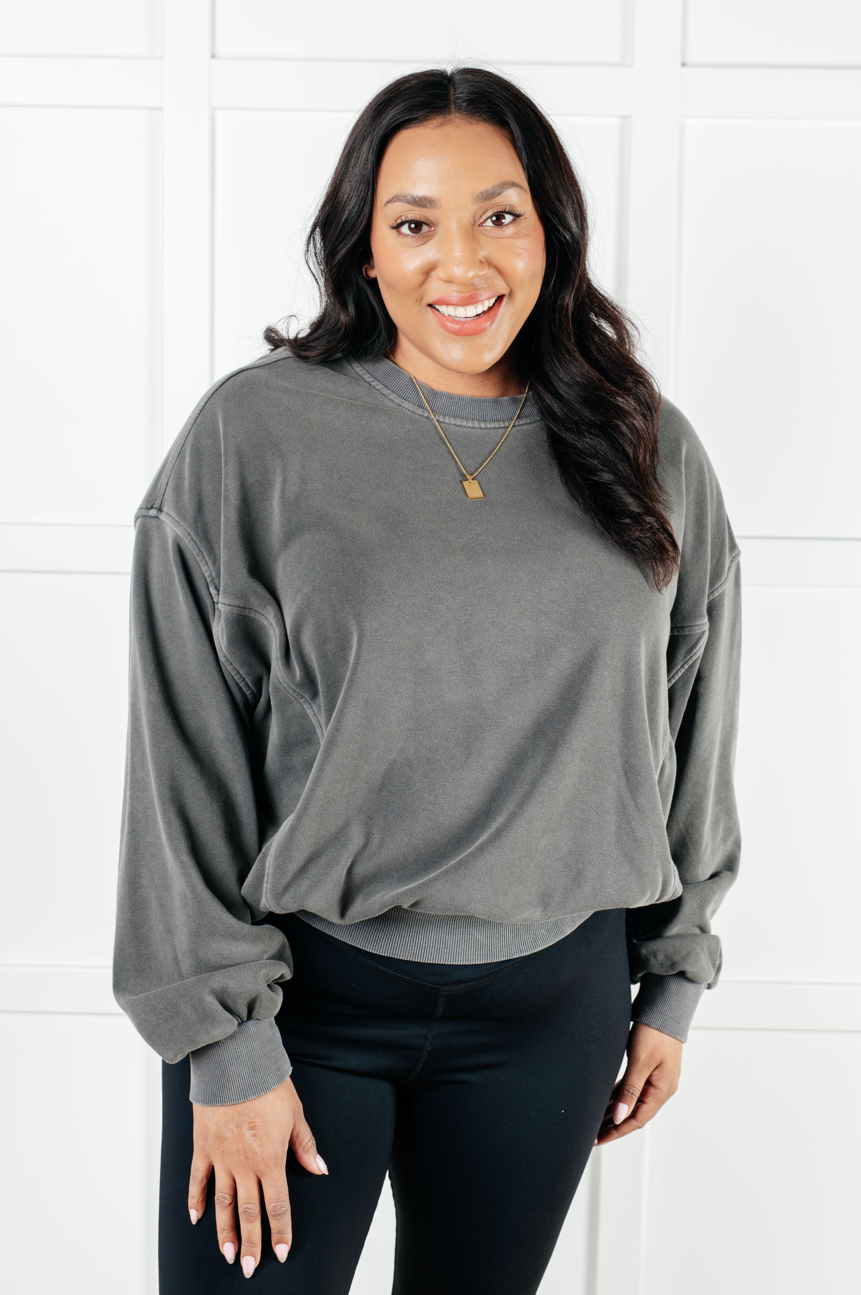 Smiling woman with dark hair wearing oversized ash black pullover and black leggings standing against white paneled wall