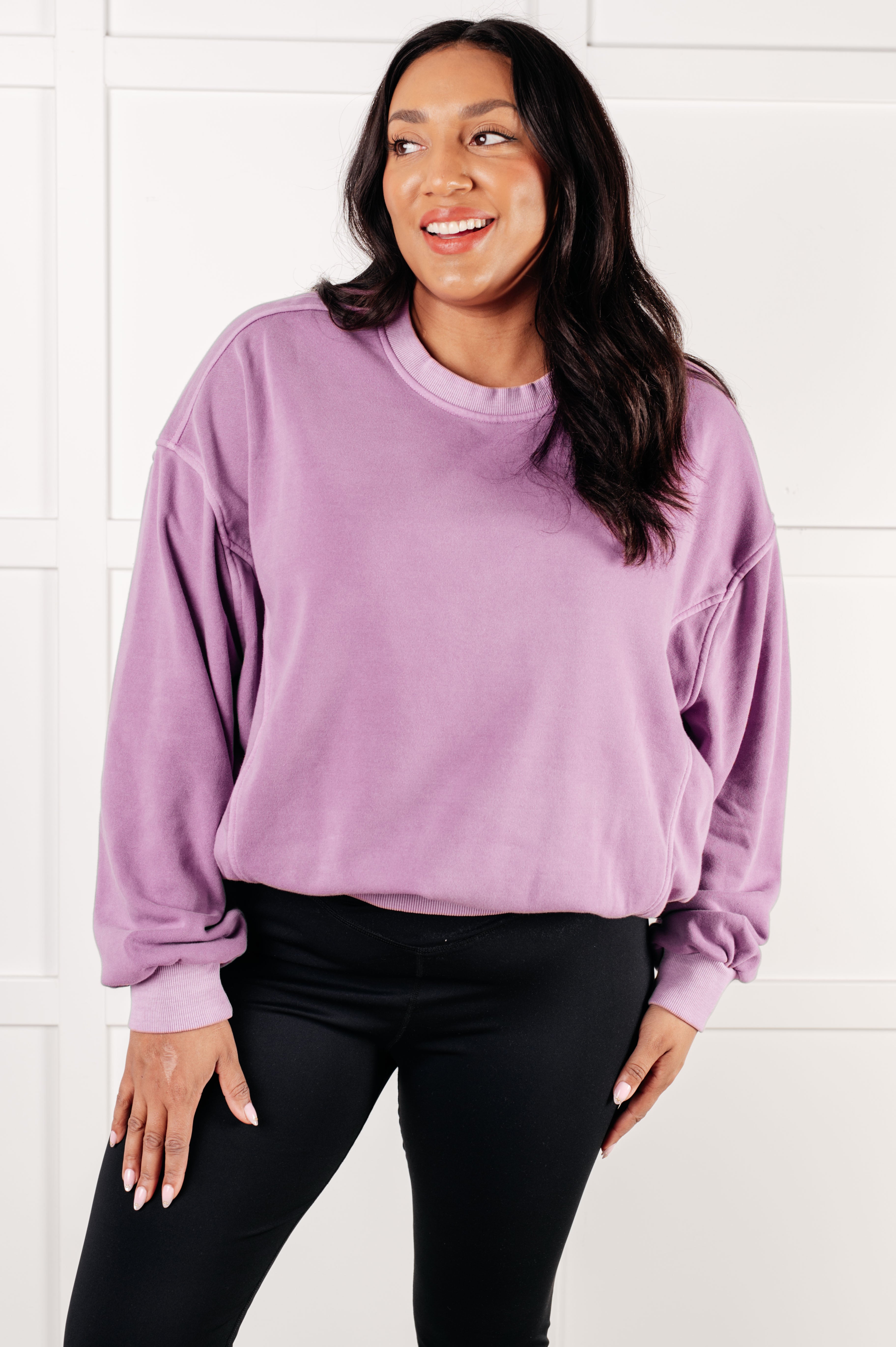 Smiling woman with long dark hair wearing oversized violet pullover and black leggings against white paneled wall
