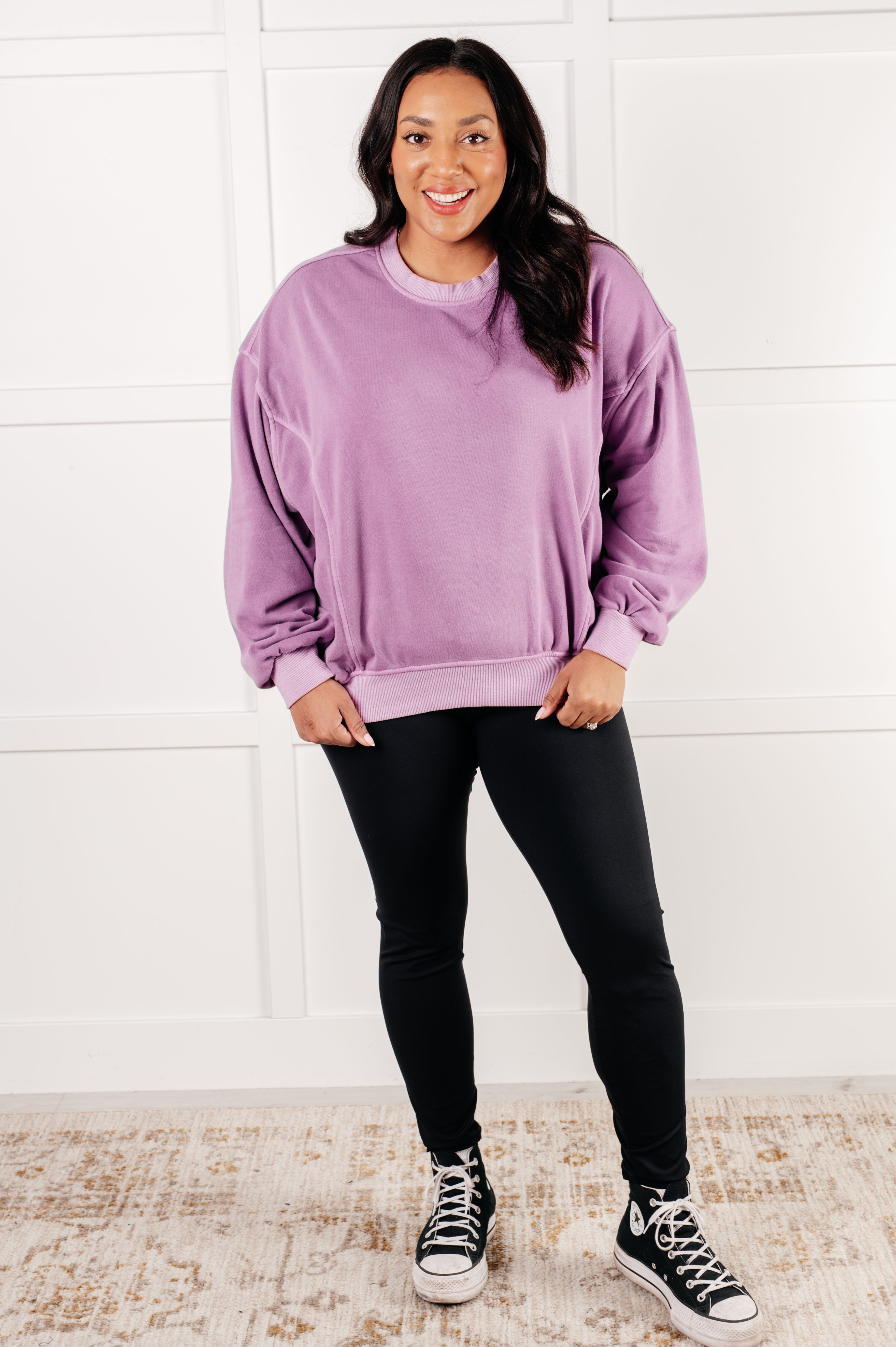 Smiling woman with long dark hair wearing relaxed violet pullover and black leggings standing on light rug against white paneled wall