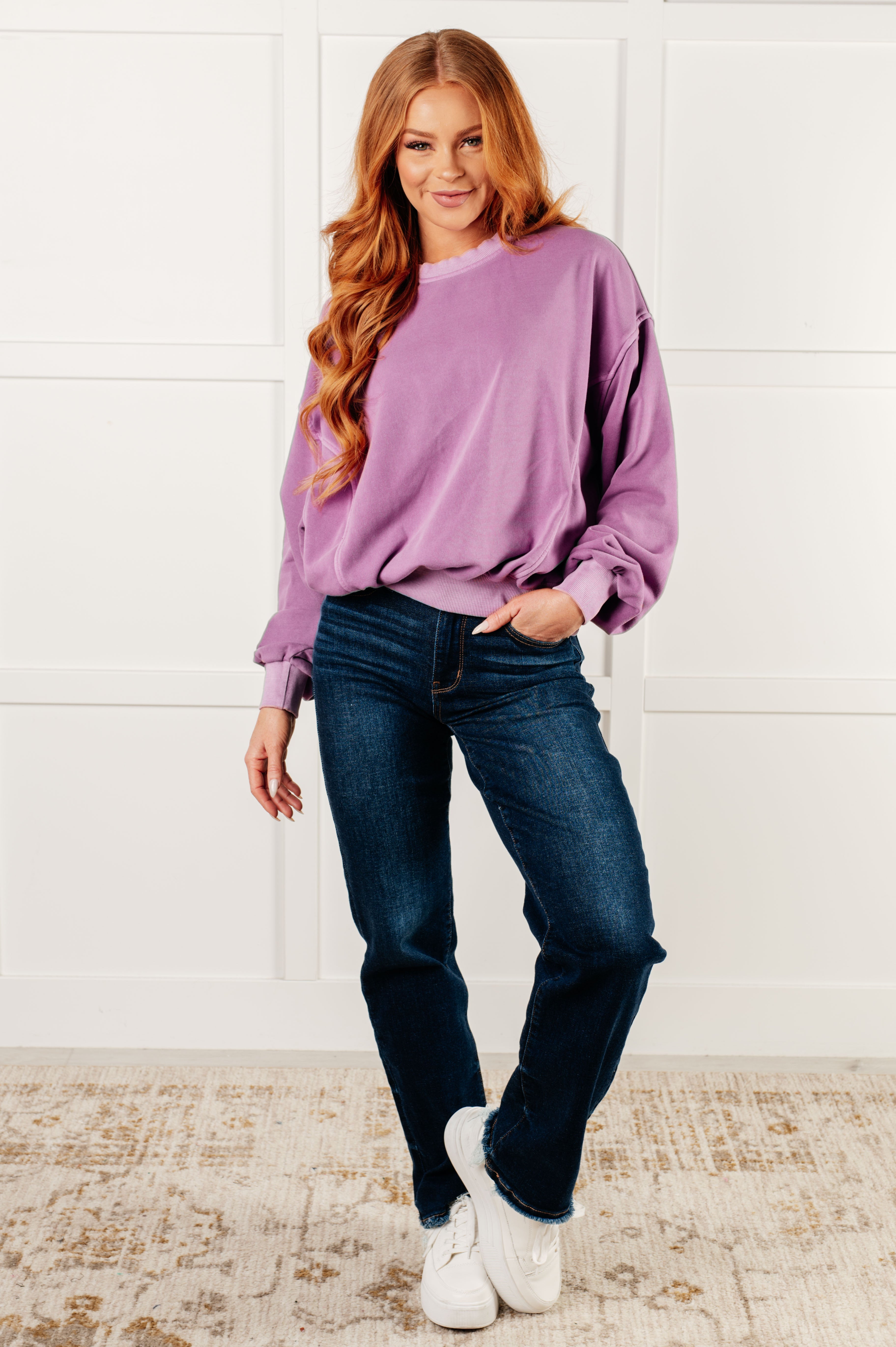Woman with long red hair wearing loose violet pullover and dark blue jeans standing on patterned rug against white paneled wall