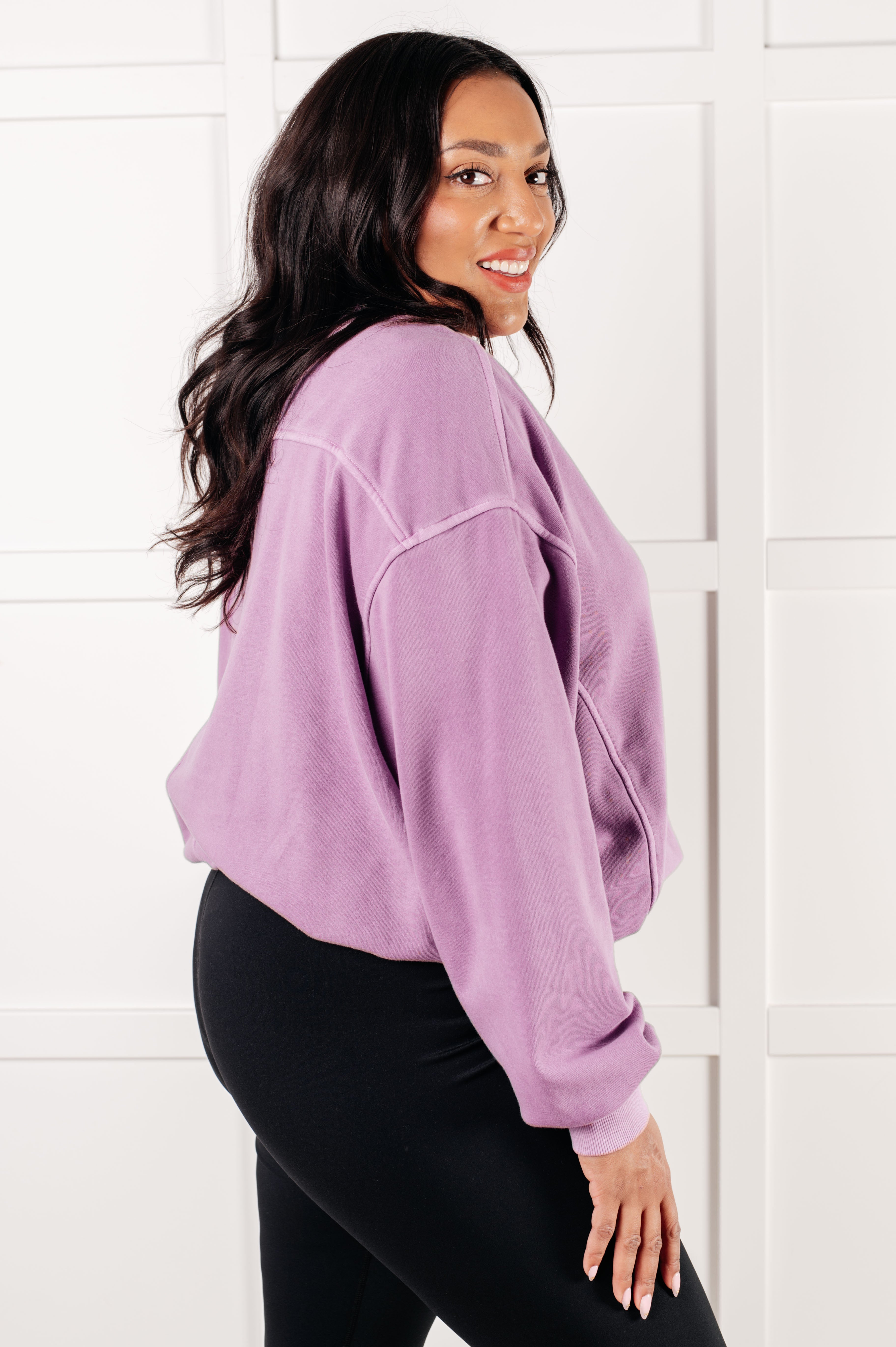 Smiling woman with long dark hair wearing violet oversized pullover and black leggings against white paneled background