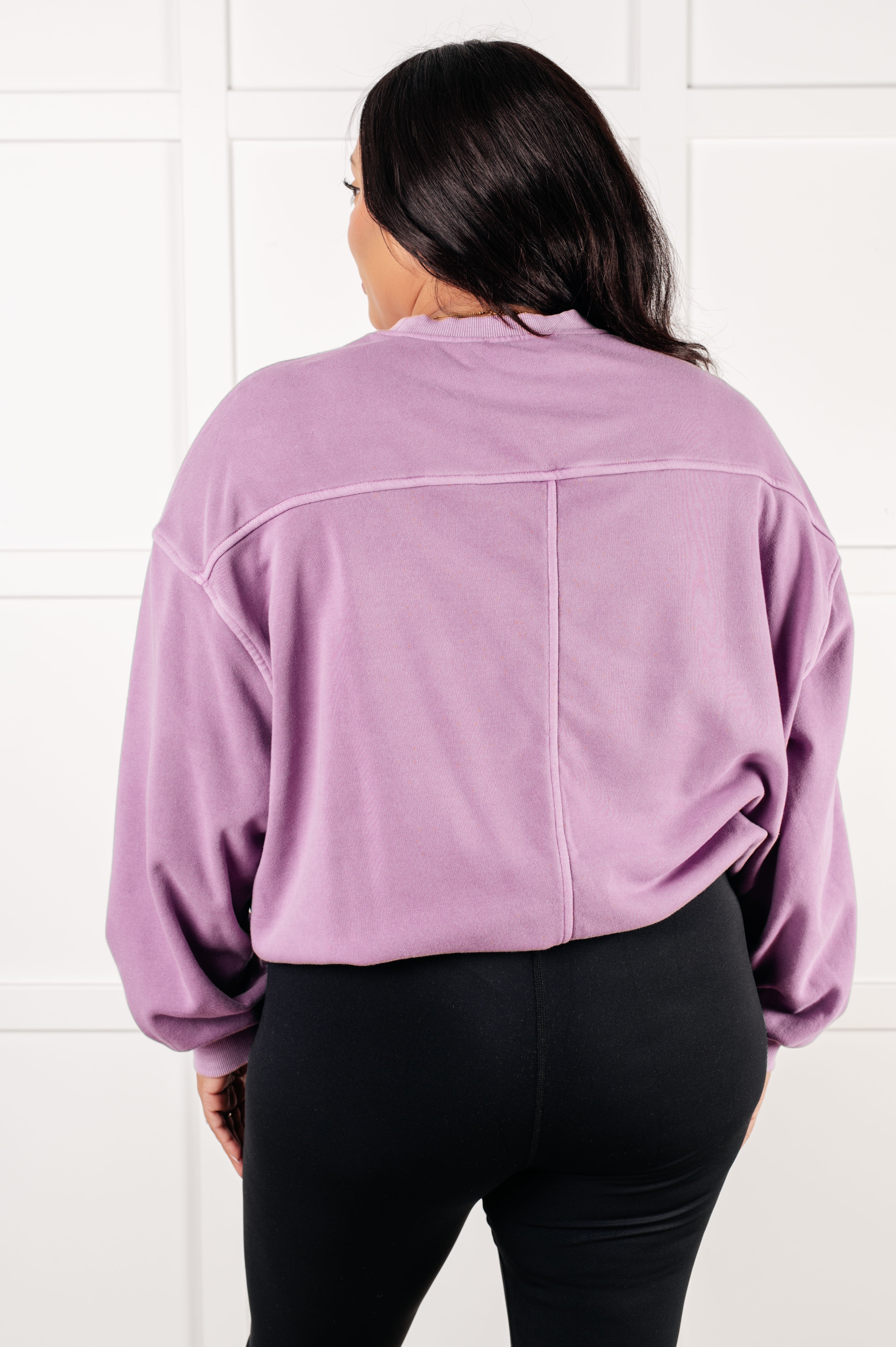 Woman with dark hair wearing loose violet pullover viewed from the back against white paneled wall