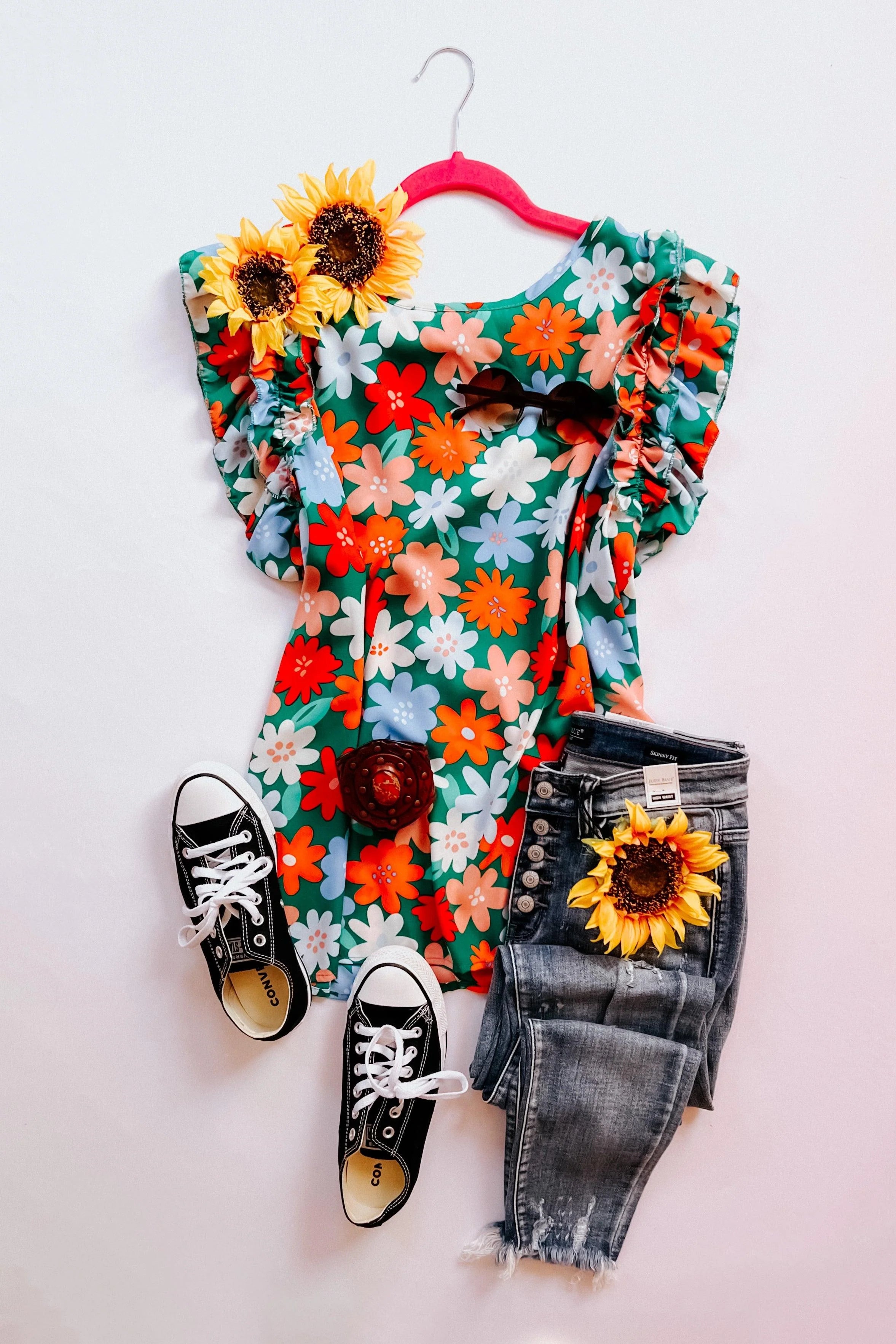 Bright floral double ruffle sleeve top on pink hanger with distressed dark denim jeans, black sneakers, and sunflowers styled flat lay