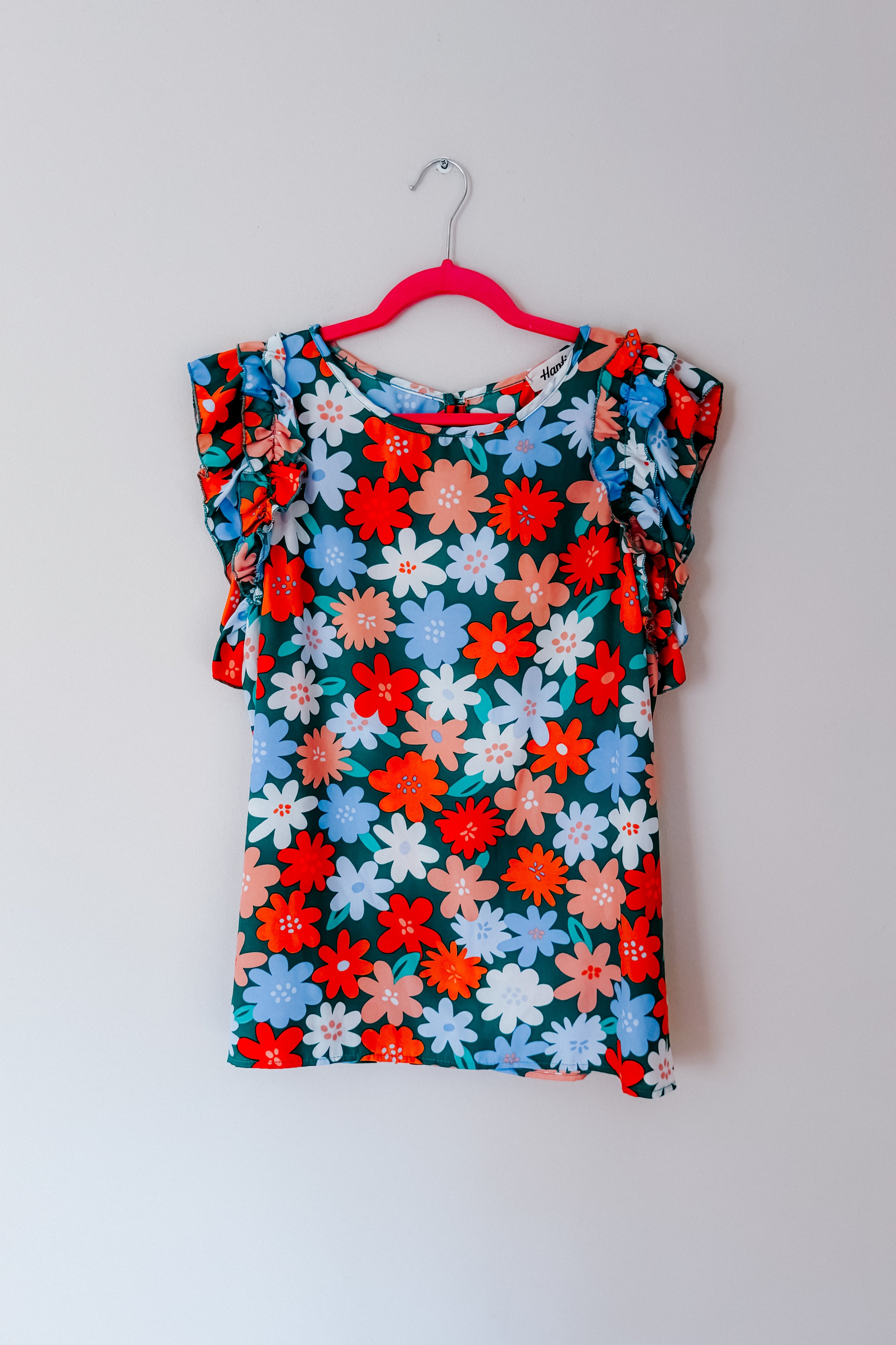 Colorful floral print sleeveless top with double ruffle sleeves hanging on pink hanger against white wall