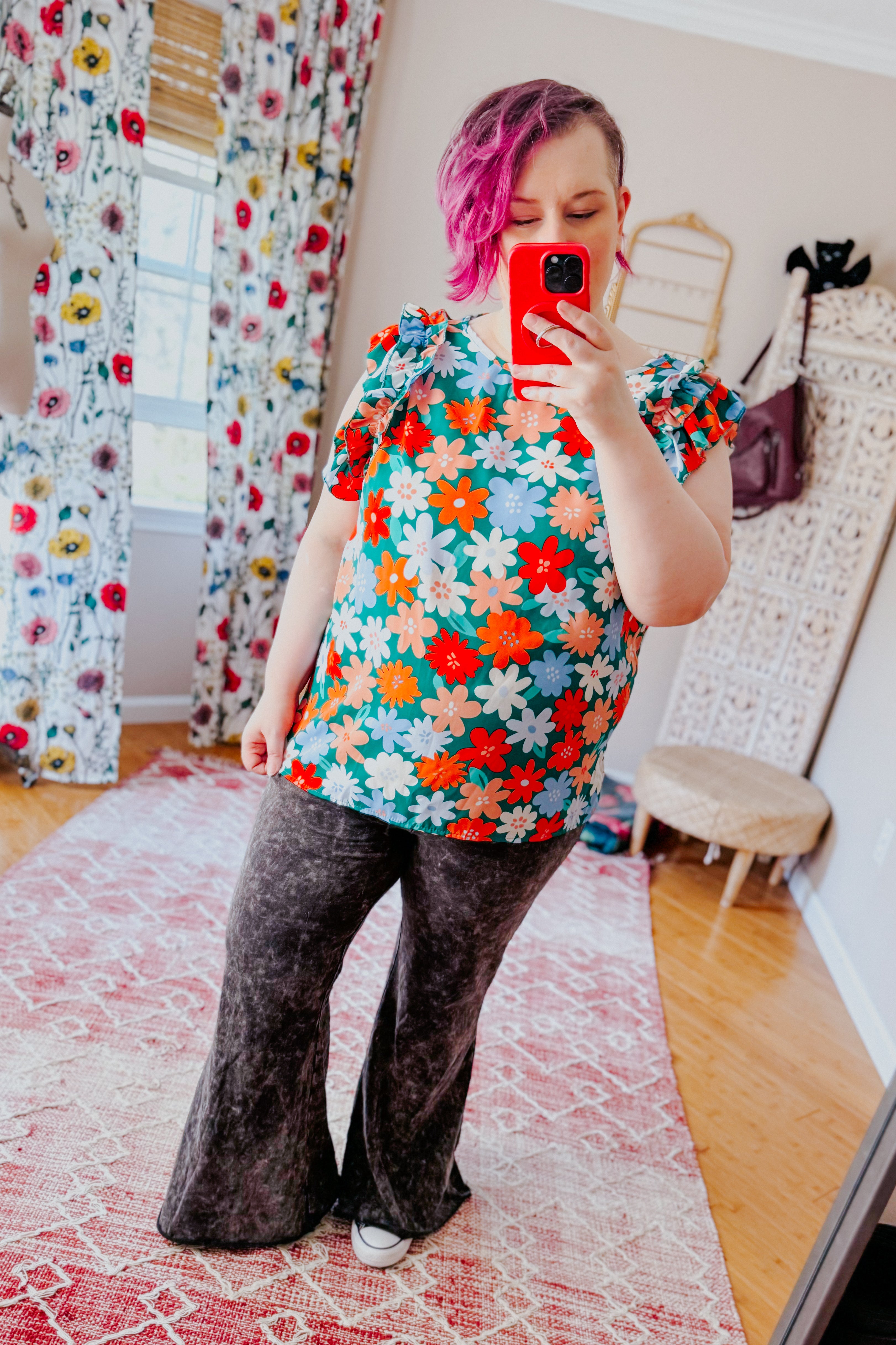 Woman with short pink hair wearing colorful floral ruffle sleeve top and dark flare pants taking mirror selfie in bright room
