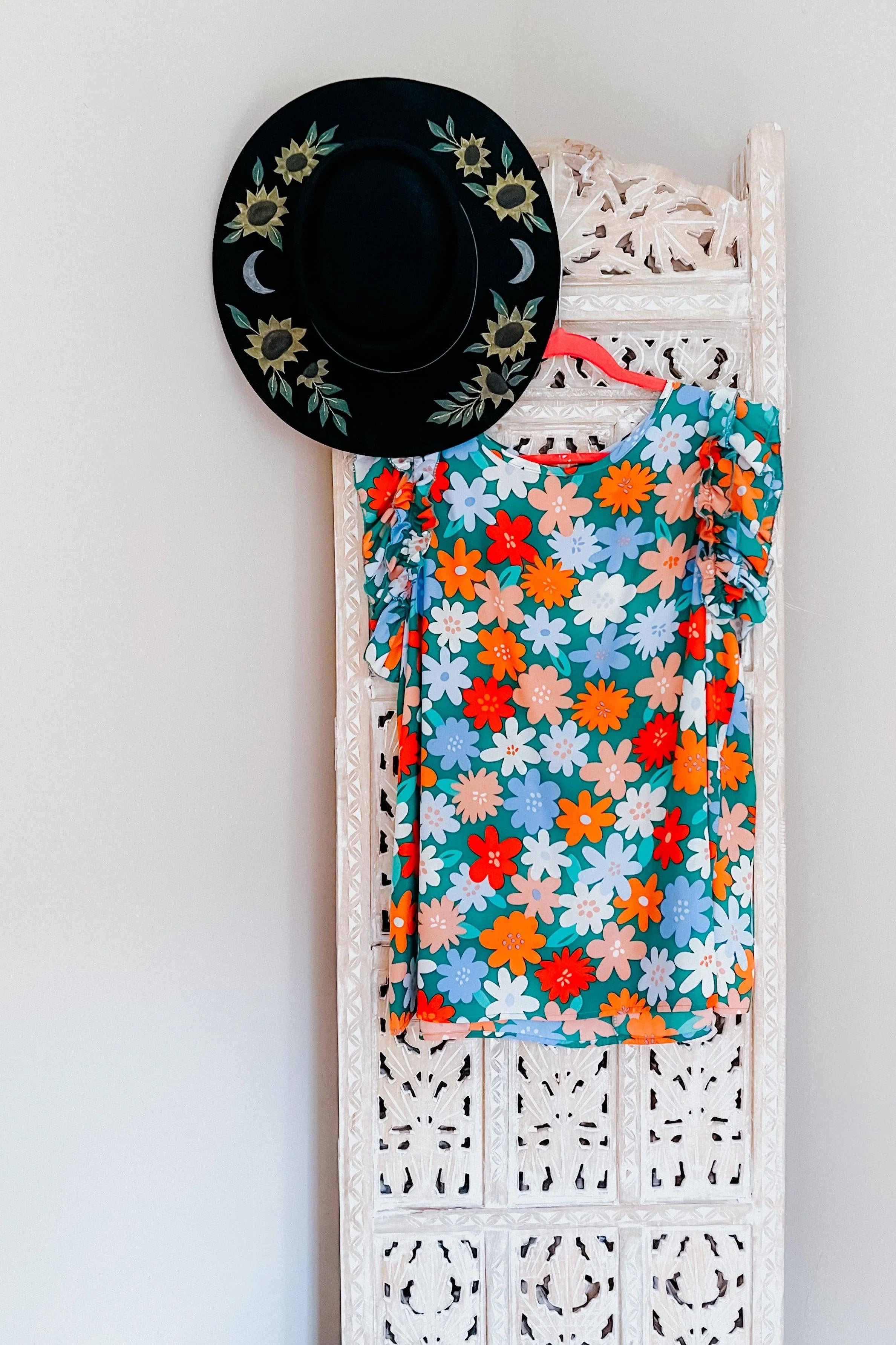 Colorful floral dress with double ruffle sleeves hanging on carved white panel next to black sunflower hat