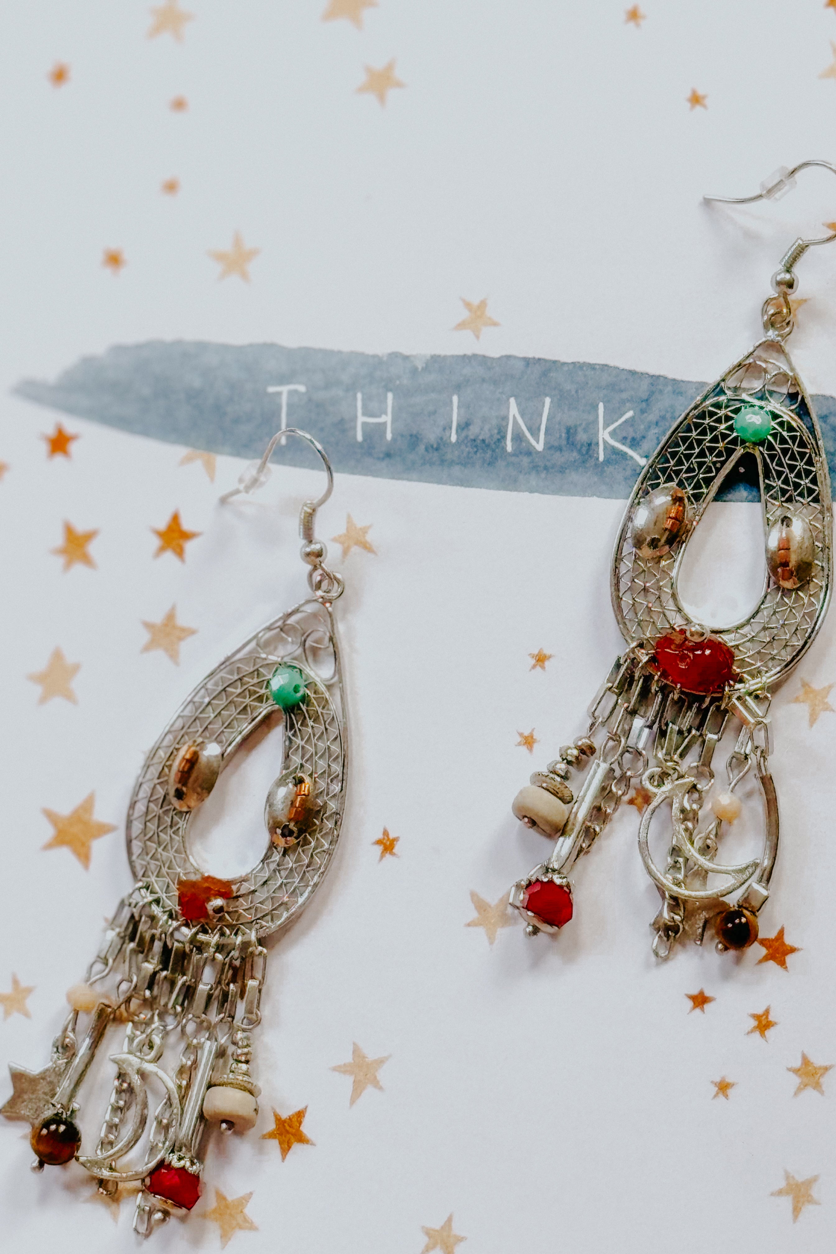 Teardrop-shaped silver earrings with moon, star charms, red and green beads on a star-patterned background with word "THINK”