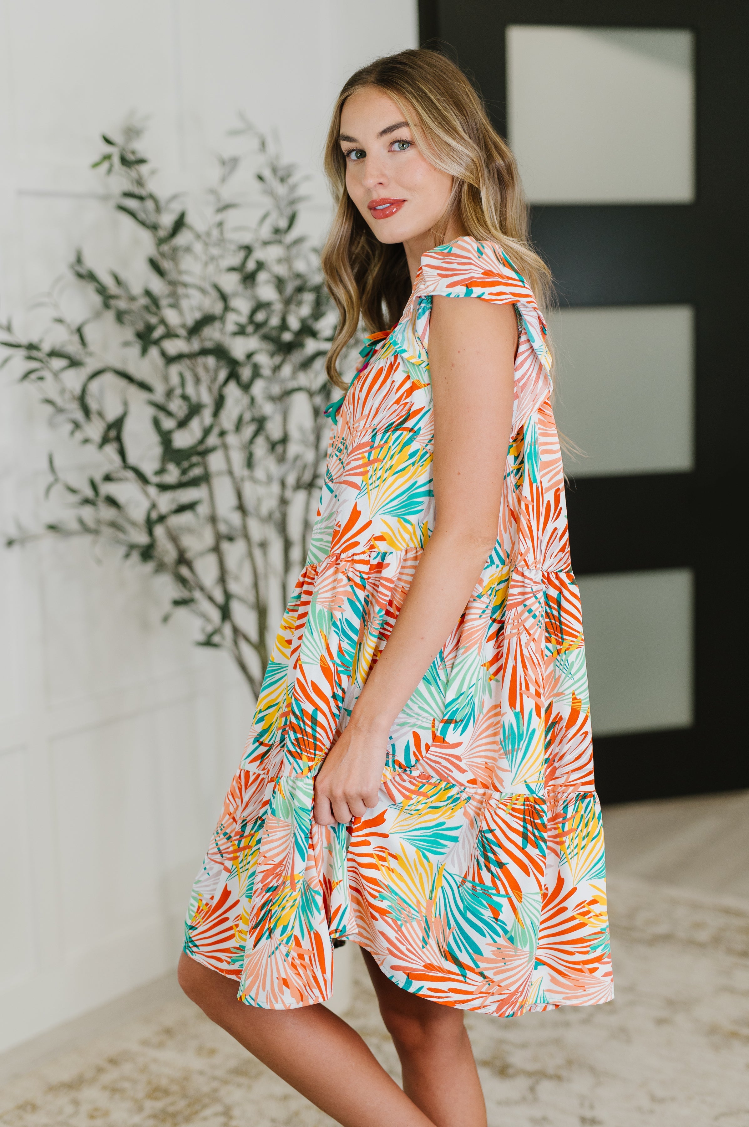 Woman wearing a colorful tiered dress with flutter sleeves in a bright indoor space with greenery background