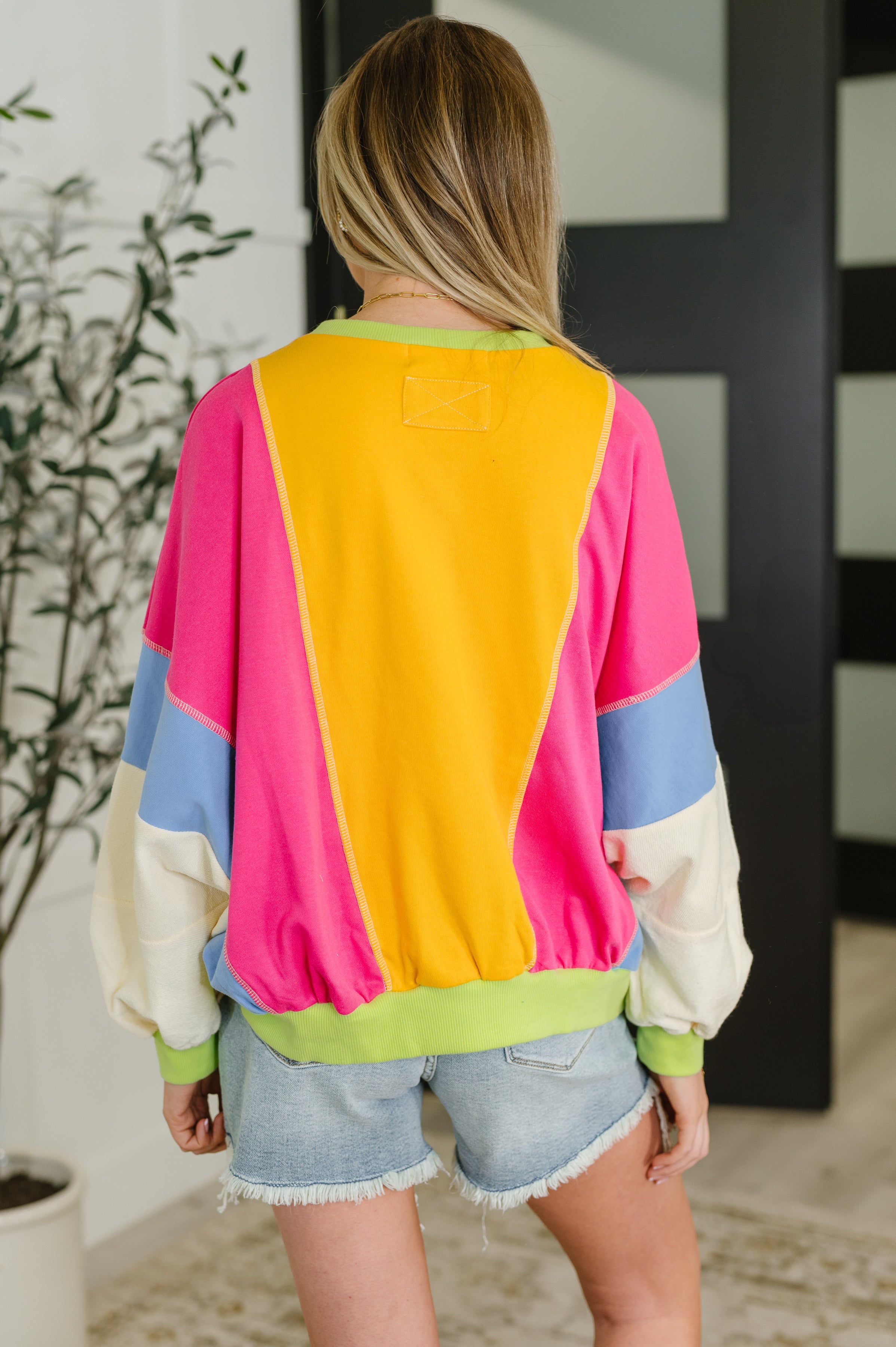 Back view of woman in a bright yellow and fuchsia color block pullover with blue and cream sleeves and denim shorts