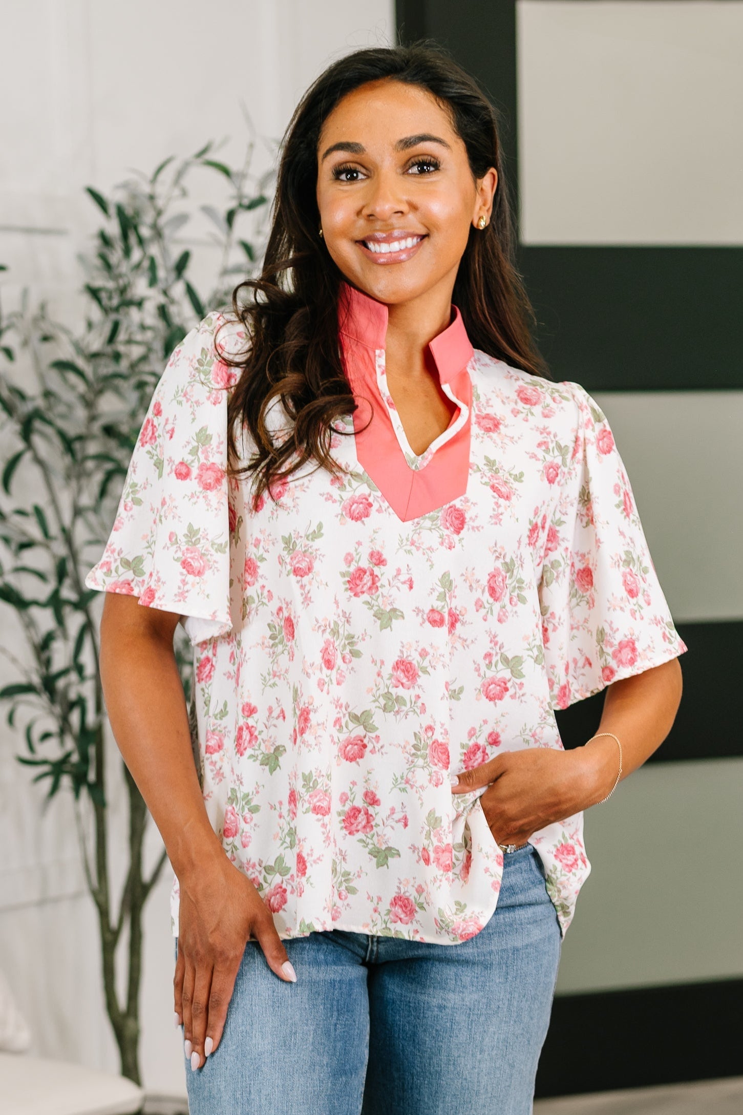 Woman wearing a coral-accented floral blouse with flutter sleeves in a bright, eclectic indoor space