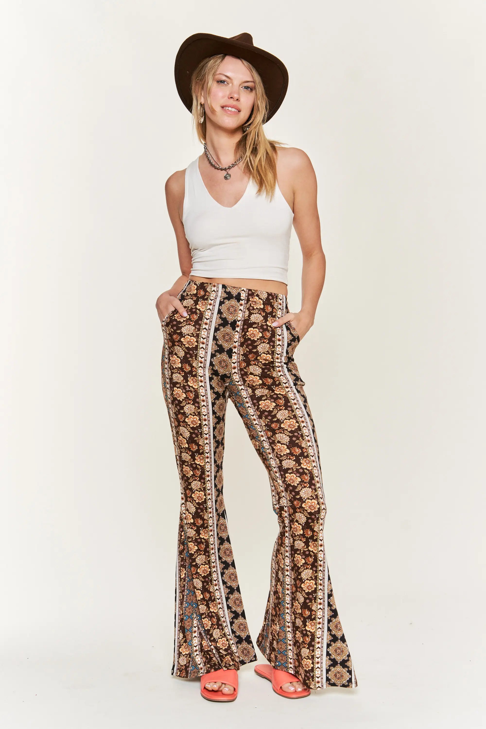 Woman wearing brown floral vertical striped flared pants with white sleeveless crop top and brown hat standing with hands in pockets