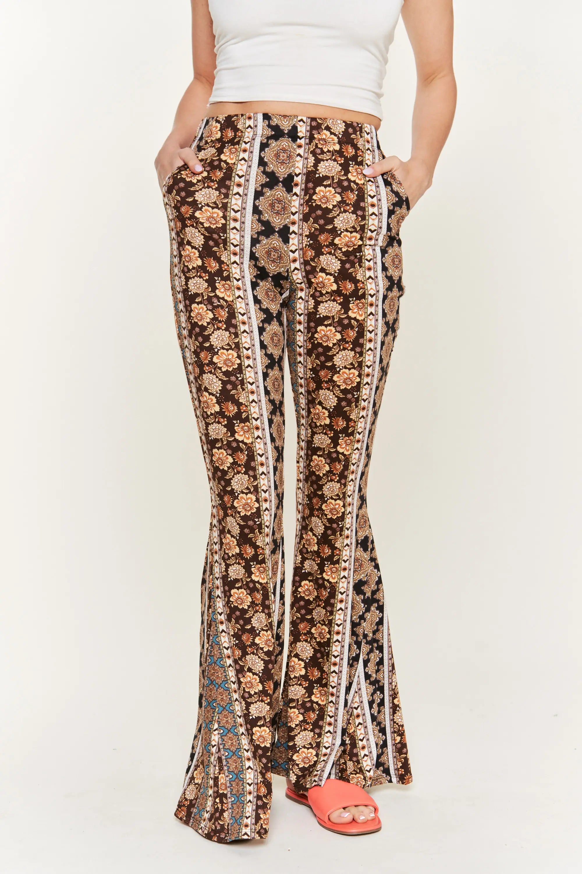 Front view of woman wearing high-waisted brown floral patterned flared pants with hands in pockets and coral slides