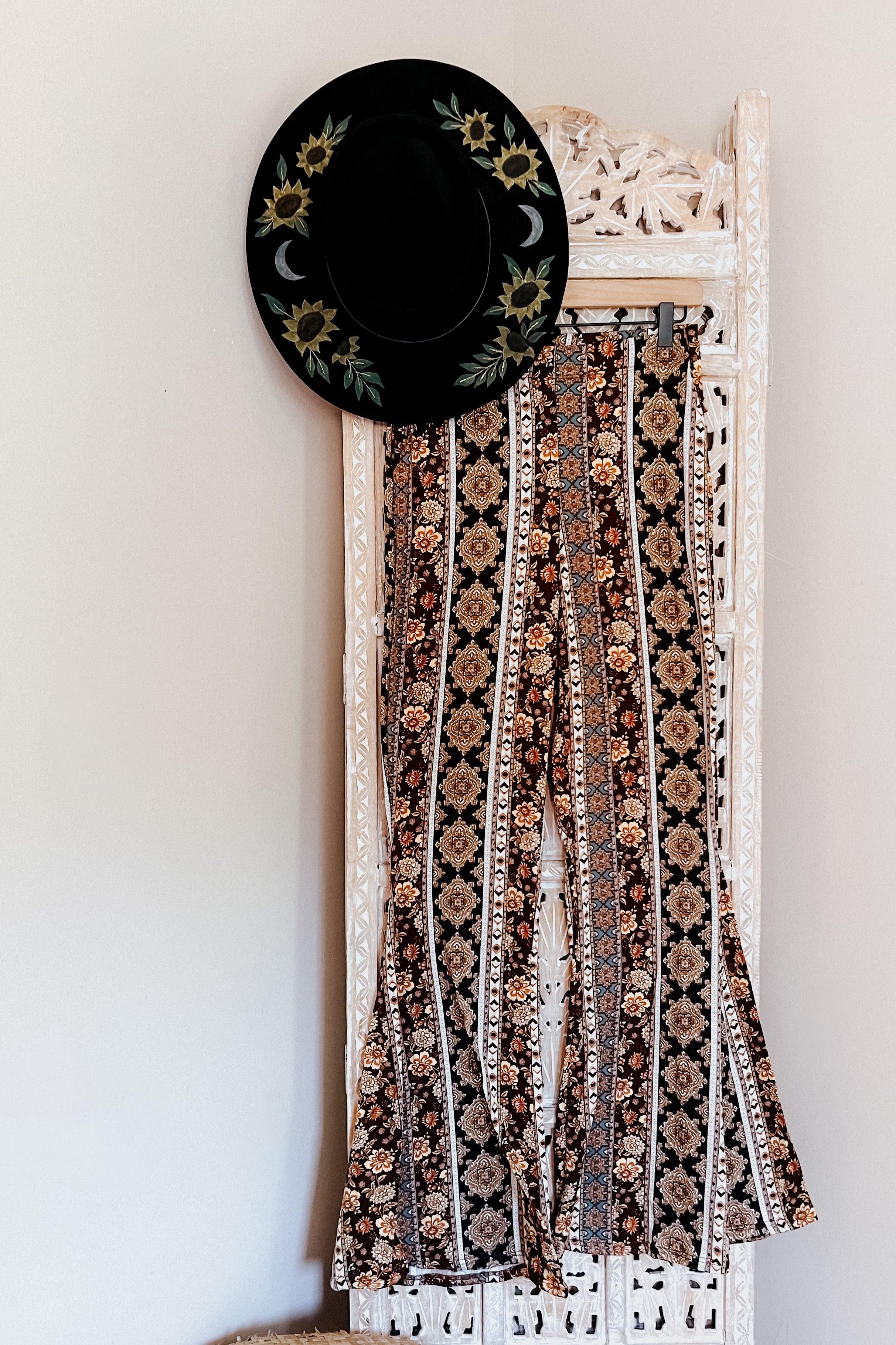 Black flared pants with intricate floral and geometric pattern hanging on white carved wooden panel with black floral hat