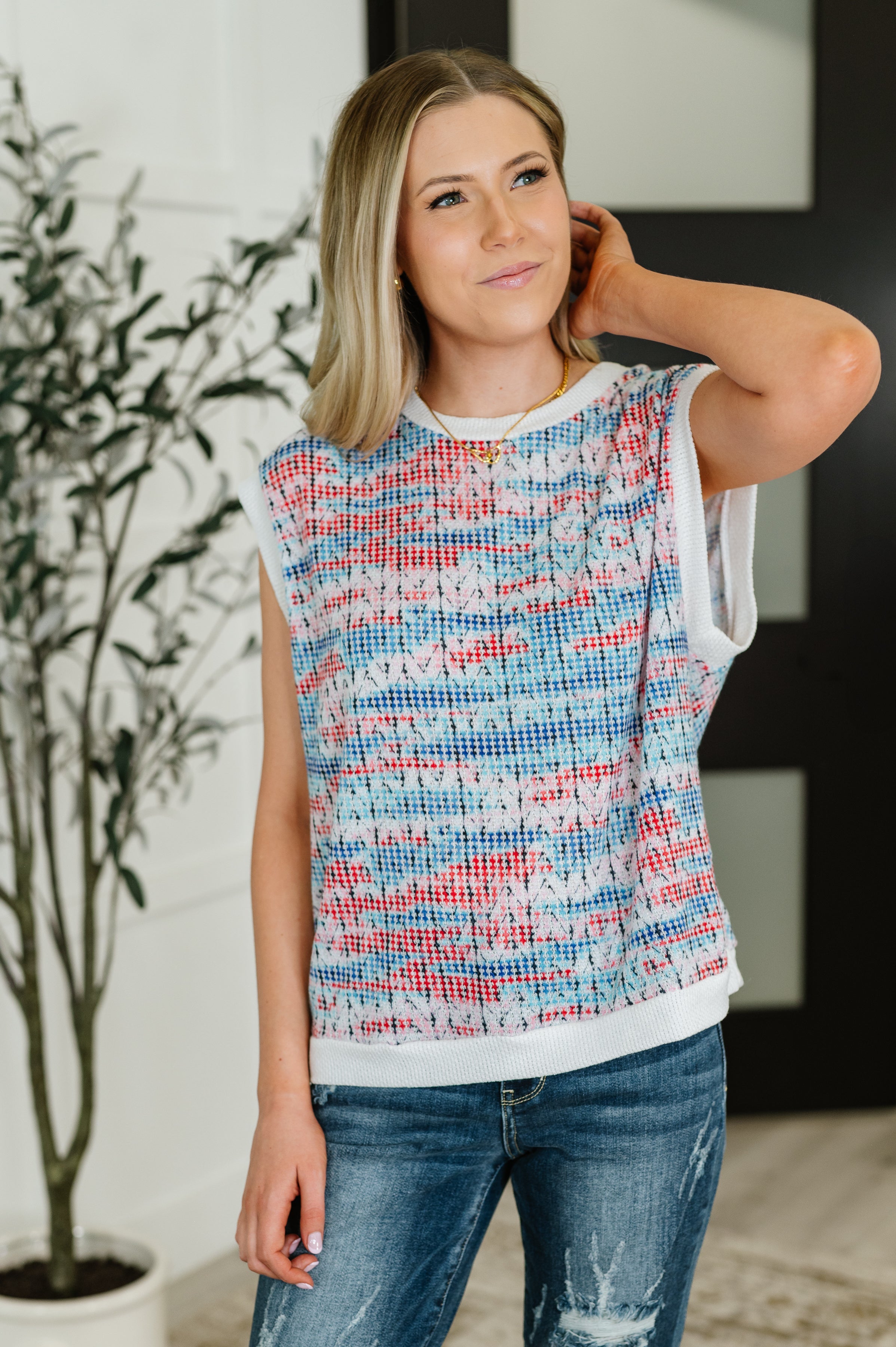 Blonde woman wearing sleeveless textured knit top with blue, red, and white abstract pattern and distressed jeans indoors